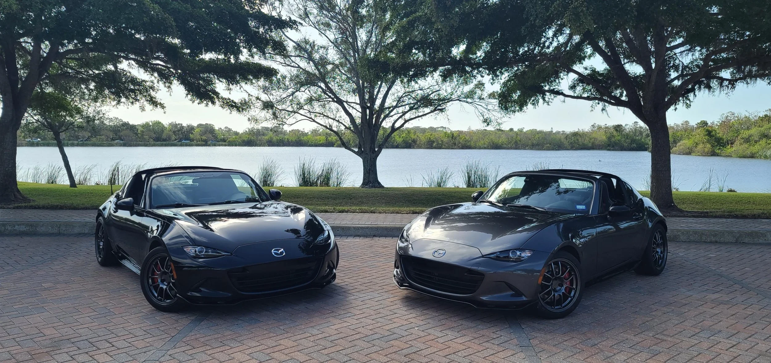 Two black Mazda sports cars parked on brick pavement by a lake with green trees in the background.