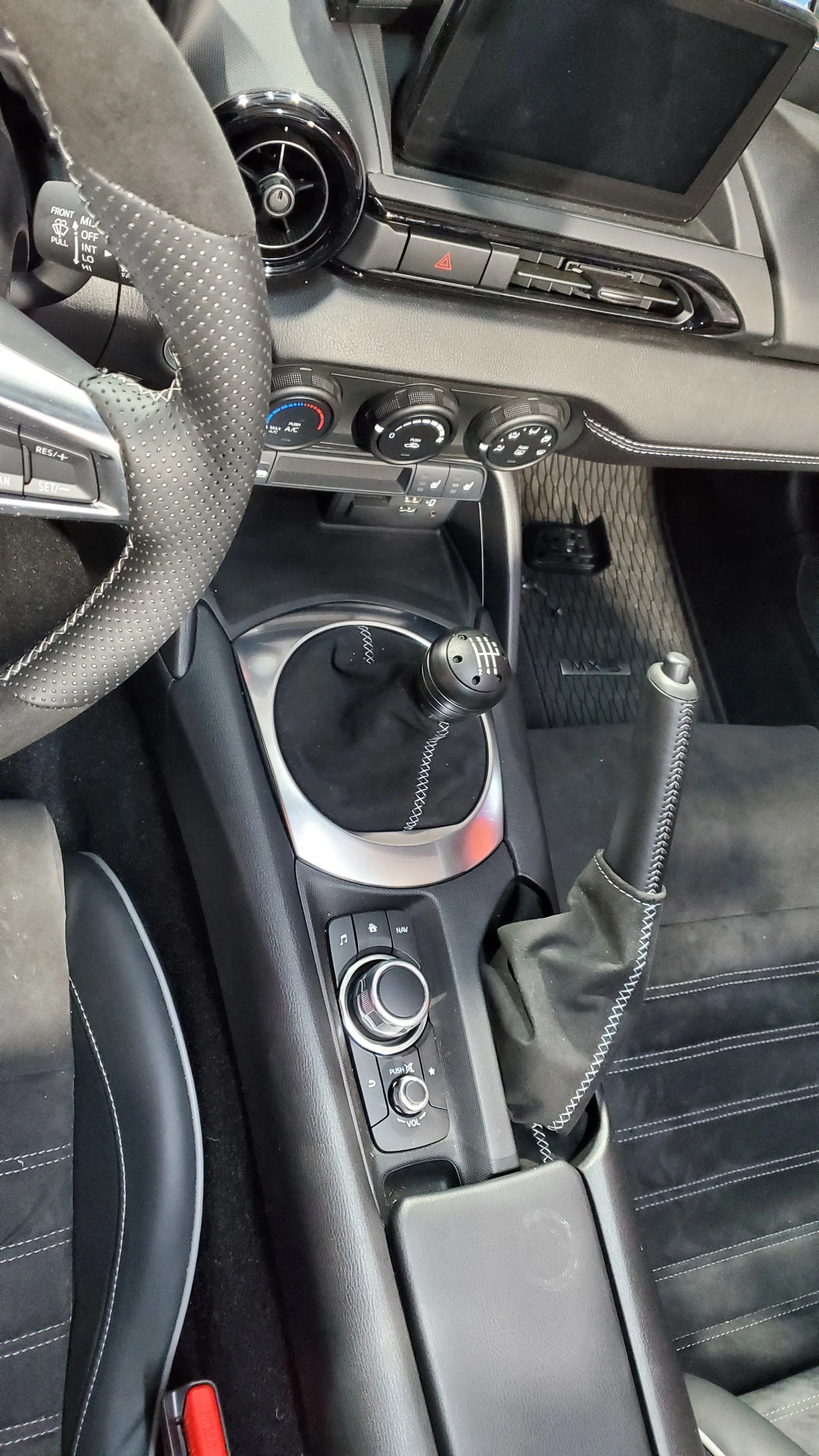 Inside view of a car's dashboard showing the steering wheel, gear shifter, handbrake, and infotainment controls with a digital display screen.