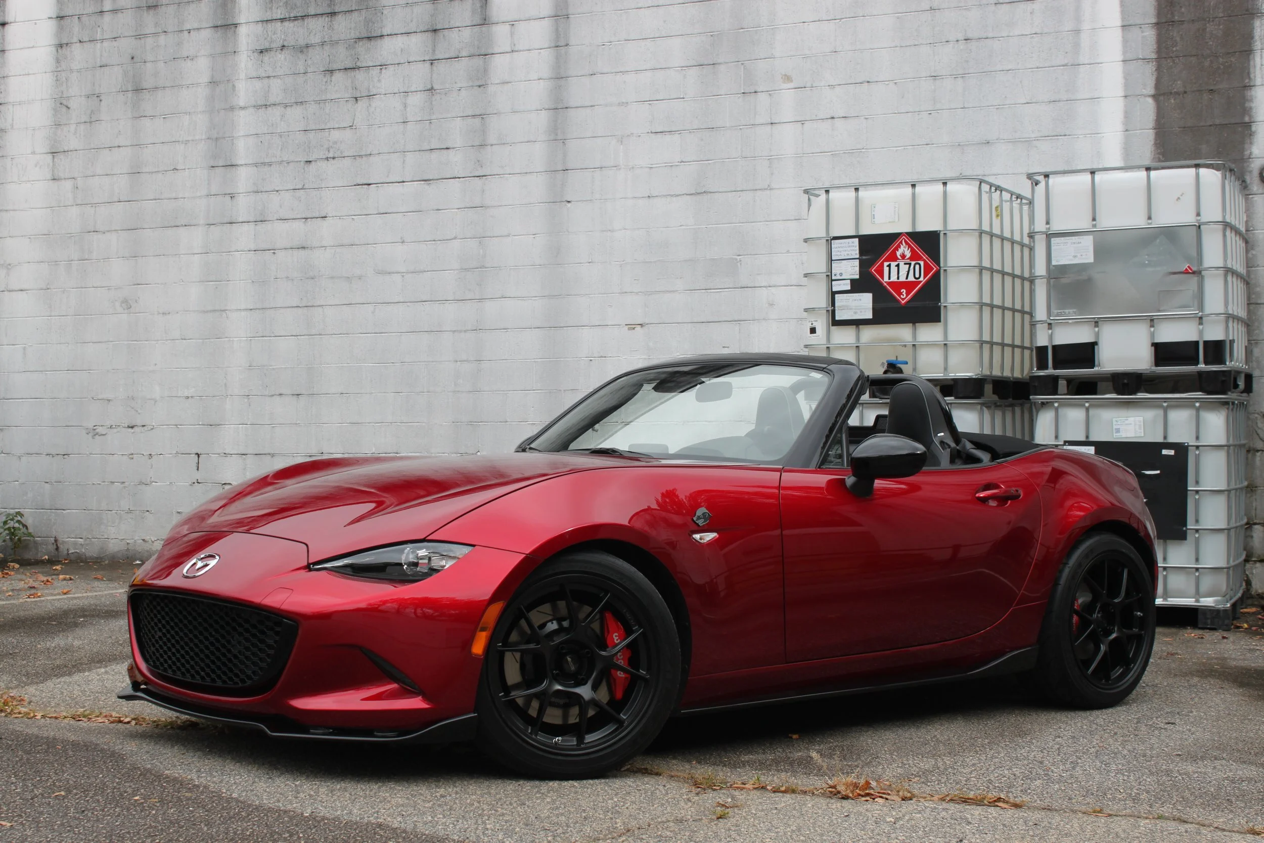 Red Mazda MX-5 convertible car parked next to a white brick wall with large industrial containers in the background.