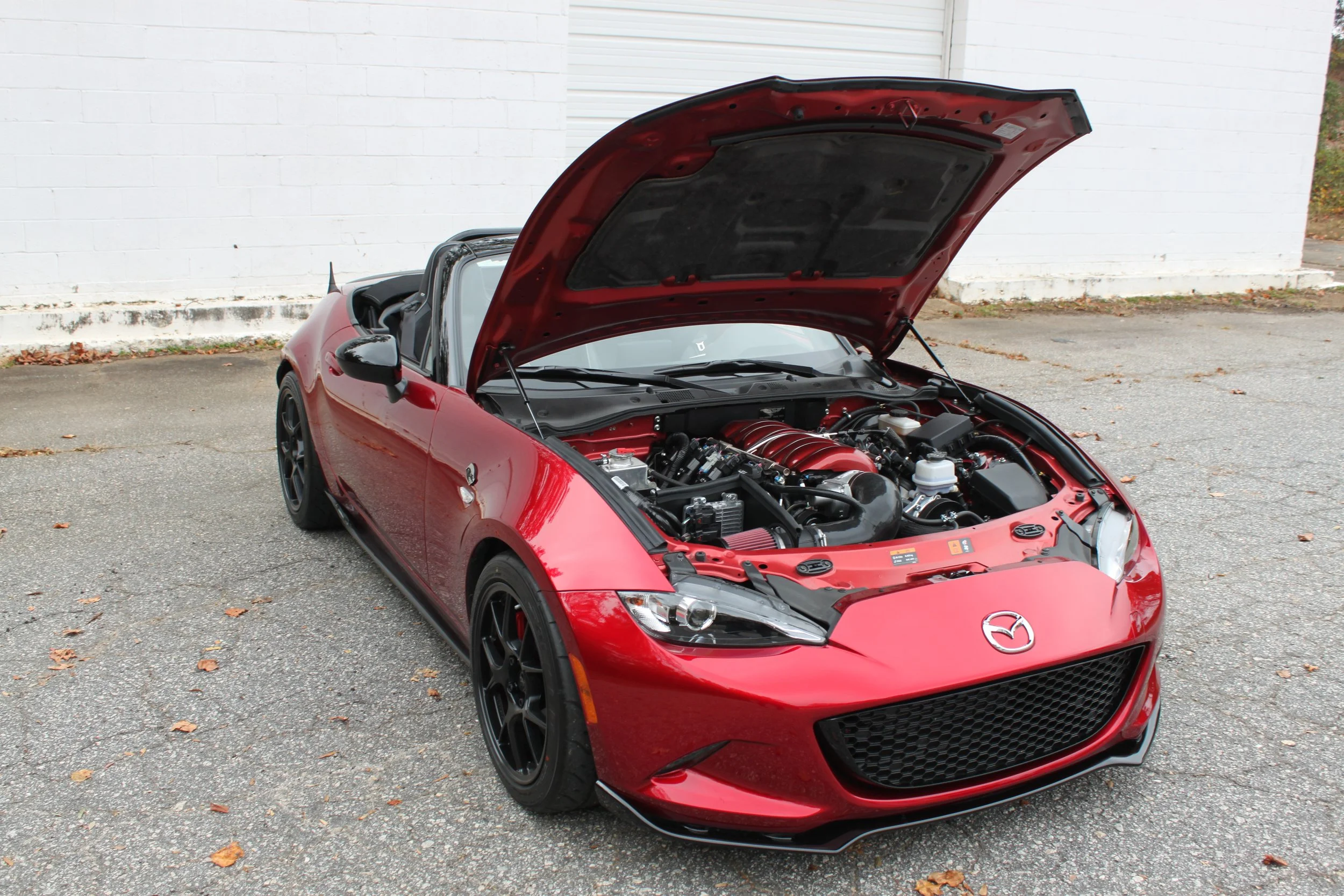 Red Mazda convertible sports car with its hood open, revealing the engine, parked on a paved surface near a white brick wall.