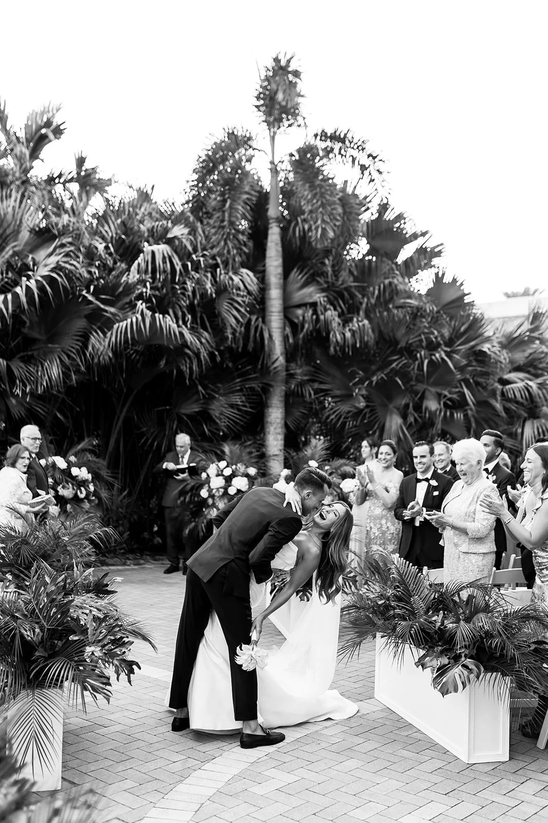 A black and white photo of a wedding ceremony outdoors with tropical plants and palm trees, showing a couple kissing, surrounded by smiling attendees.