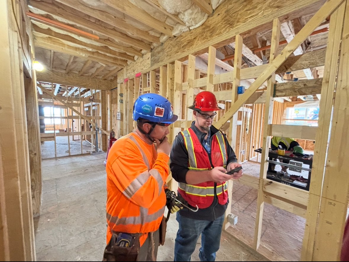 Two construction workers inside a partially framed building, wearing hard hats and high-visibility vests, discussing safety and compliance on the jobsite
