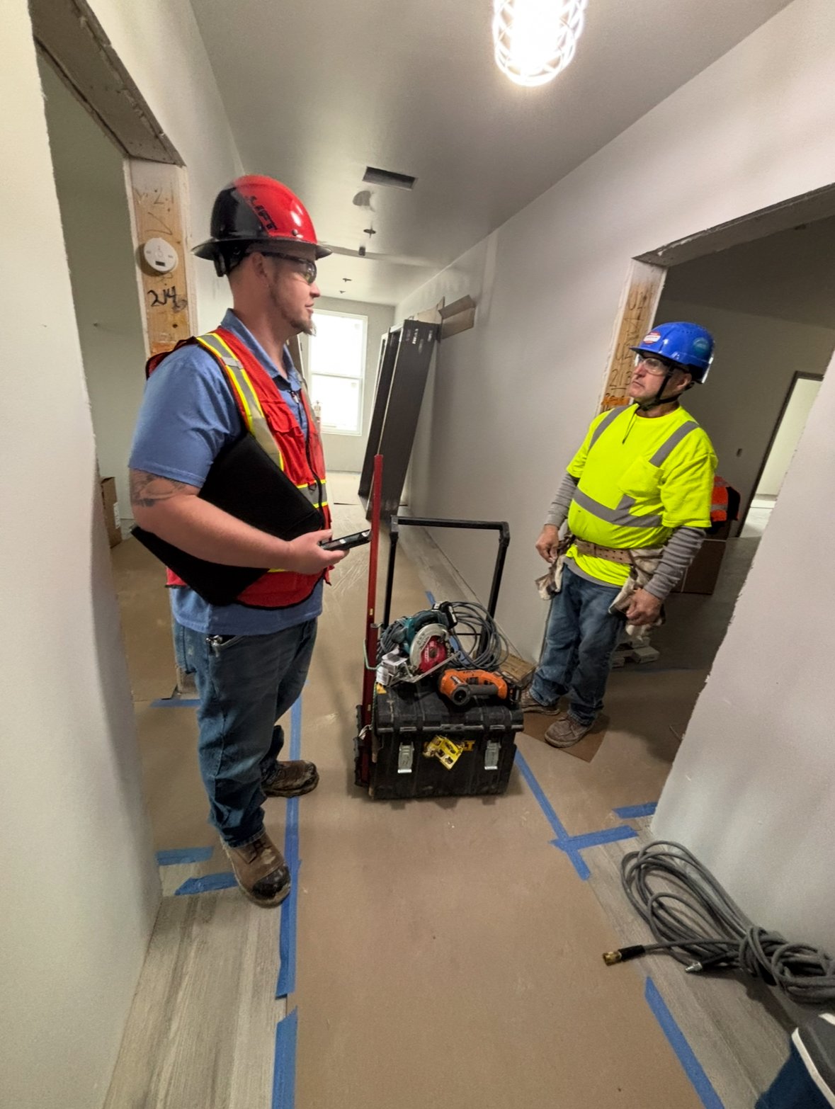 Two construction workers wearing safety helmets and high-visibility vests standing inside a house under renovation. They are discussing safety and compliance on the jobsite.