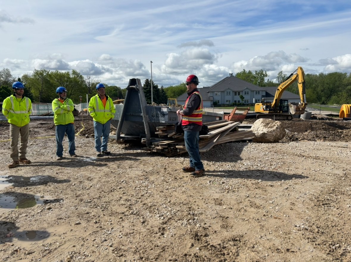 Construction workers at a site with machinery, gravel, and a large rock, under a partly cloudy sky.