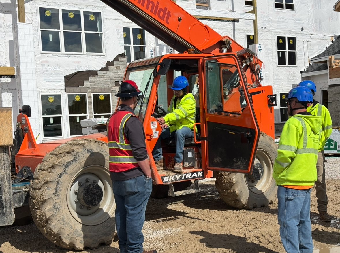 Construction workers at a building site with a SkyTrak telescopic forklift, one worker seated inside operating, others standing nearby wearing safety helmets and high-visibility vests during forklift training