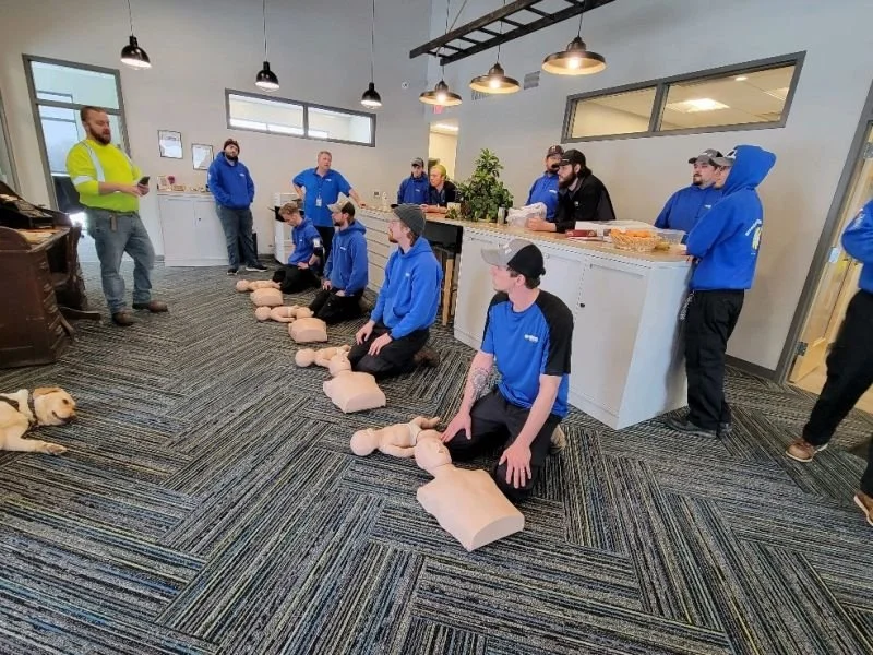 Group of people participating in a CPR training session using mannequins, with instructor and a dog present in the room.