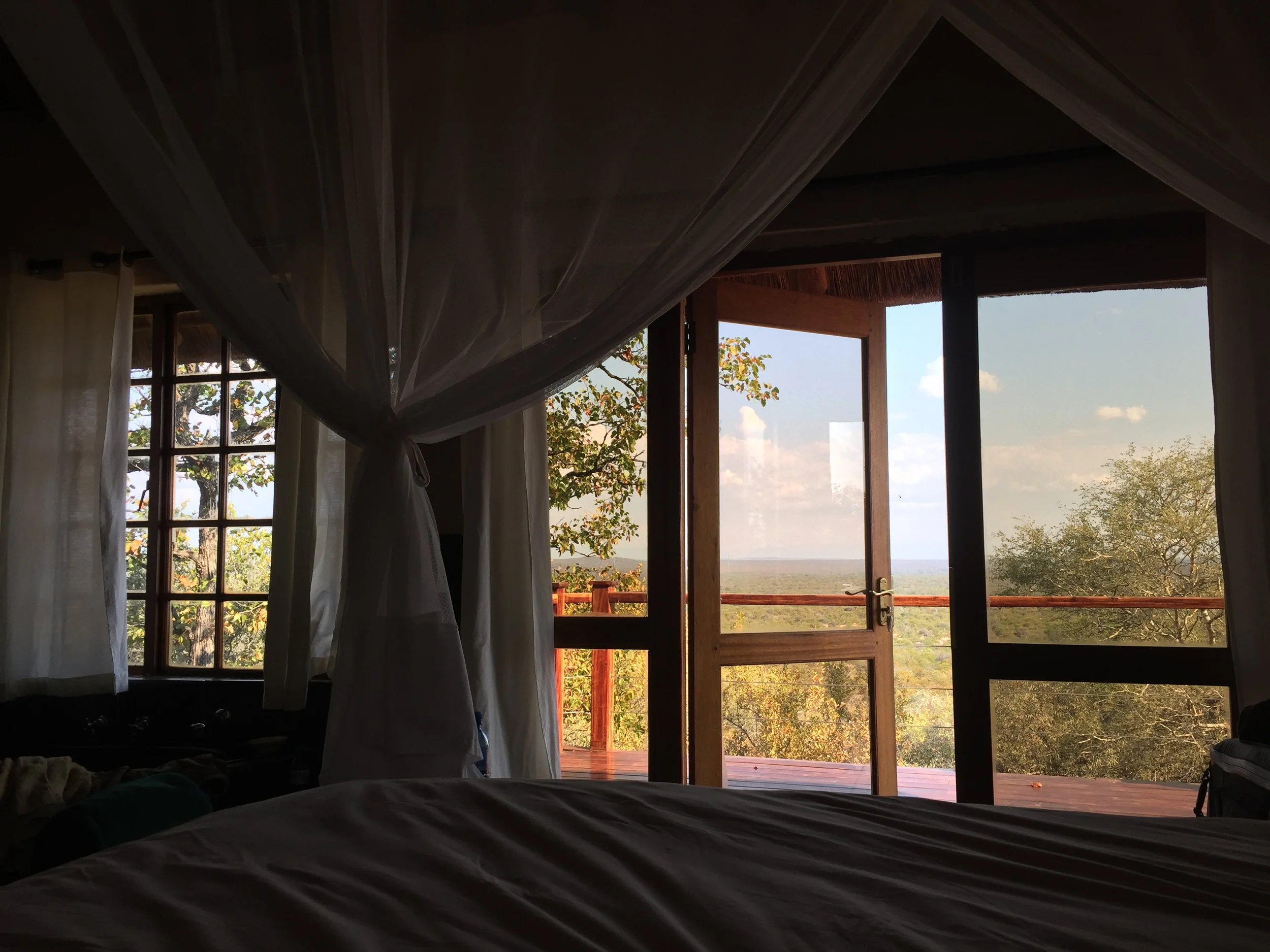 Bedroom interior with canopy bed and open doors leading to a balcony with a view of trees and a distant landscape under a blue sky.