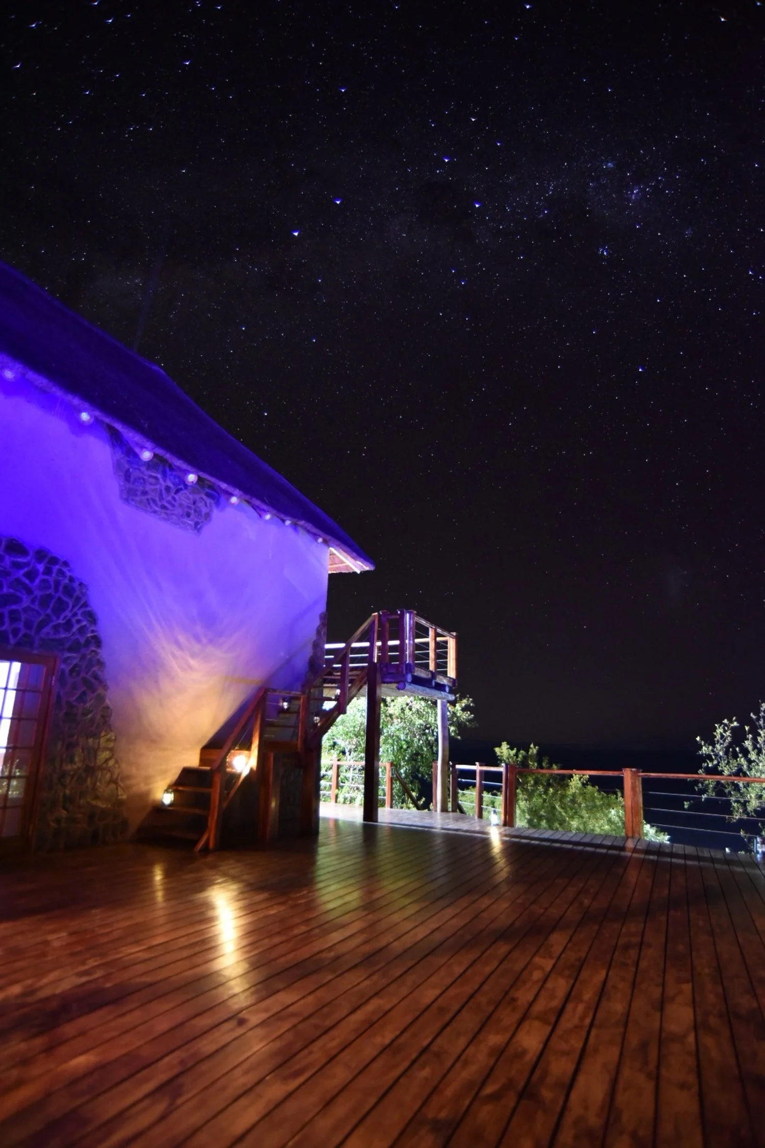 Wooden deck under a starry night sky, near a lit rustic building with stone walls and a thatched roof, featuring an outdoor staircase and railing.