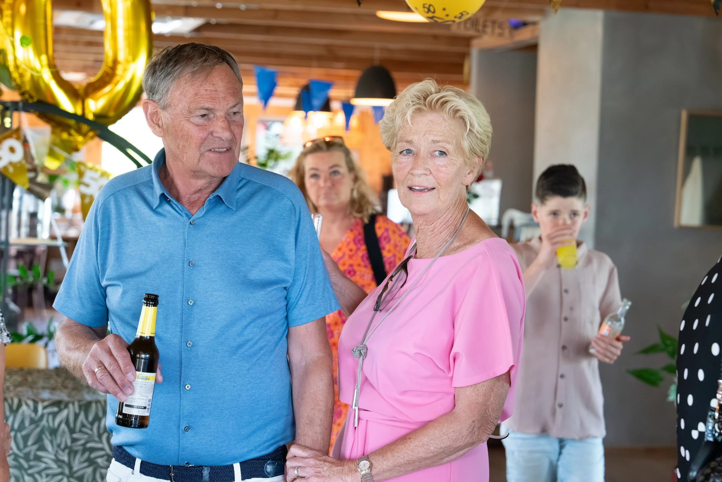 Man en vrouw die hand in hand staan tijdens een feestje, omringd door andere gasten die drinken en feestversiering in een brasserie