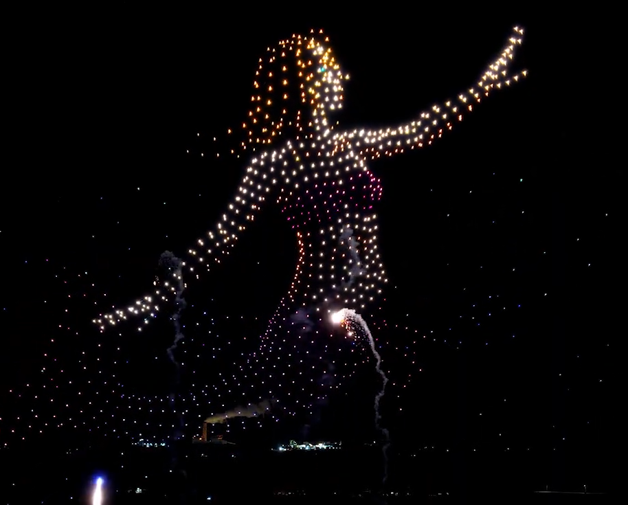 A drone show with fireworks depicting a mermaid in the sky above spectators.