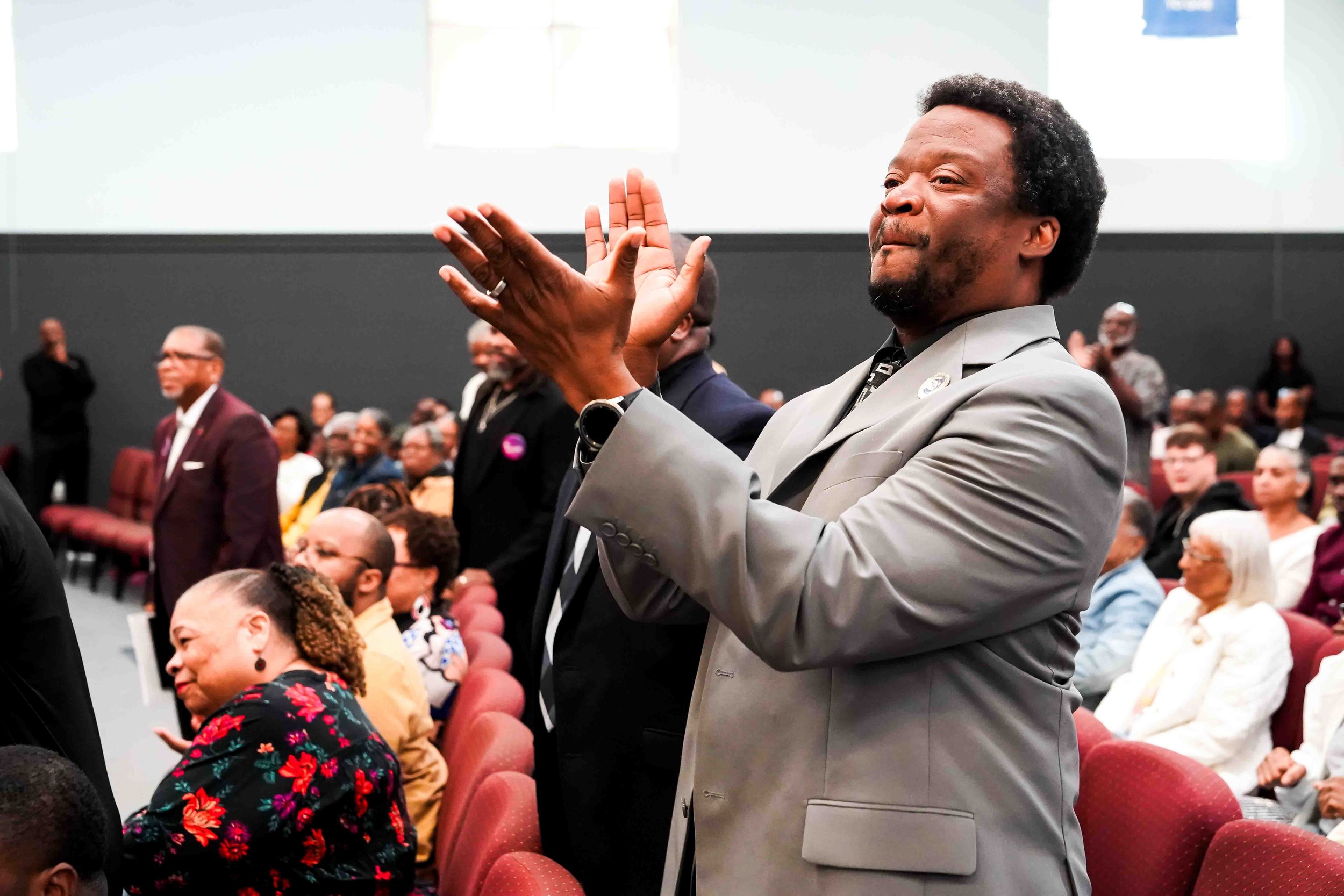 A man in a gray suit claps in a crowded auditorium during a formal event, with seated and standing attendees in the background.