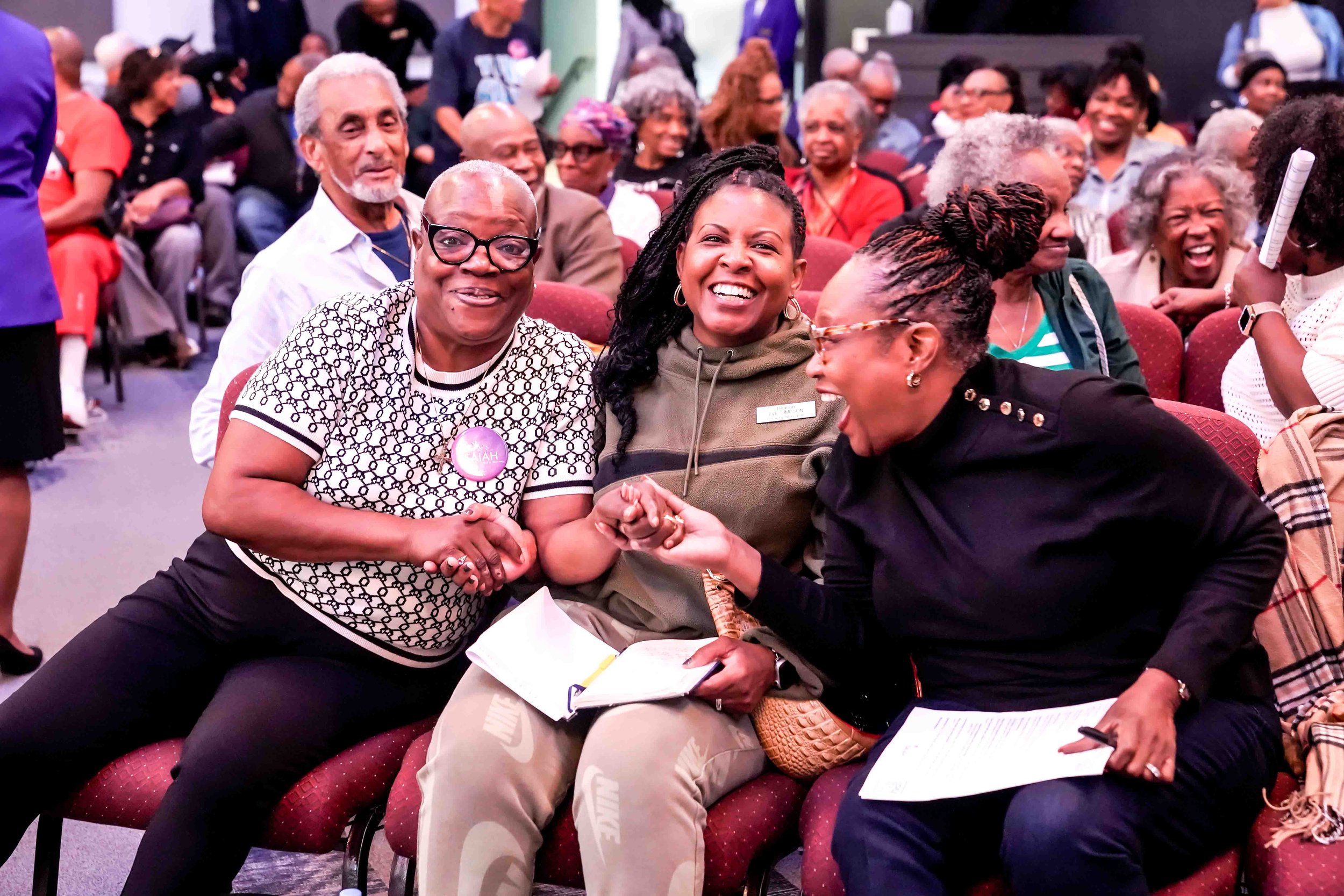 A group of Black women and men smiling and laughing at a crowded event, engaging in a friendly hand-hold handshake among three women seated in the foreground.