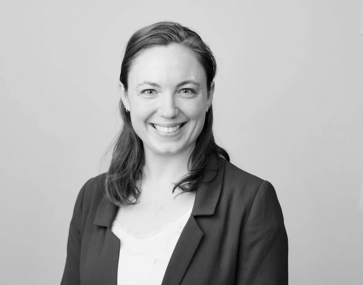Black-and-white portrait of a smiling woman with shoulder-length hair, wearing a blazer over a light-colored top, standing against a plain background.