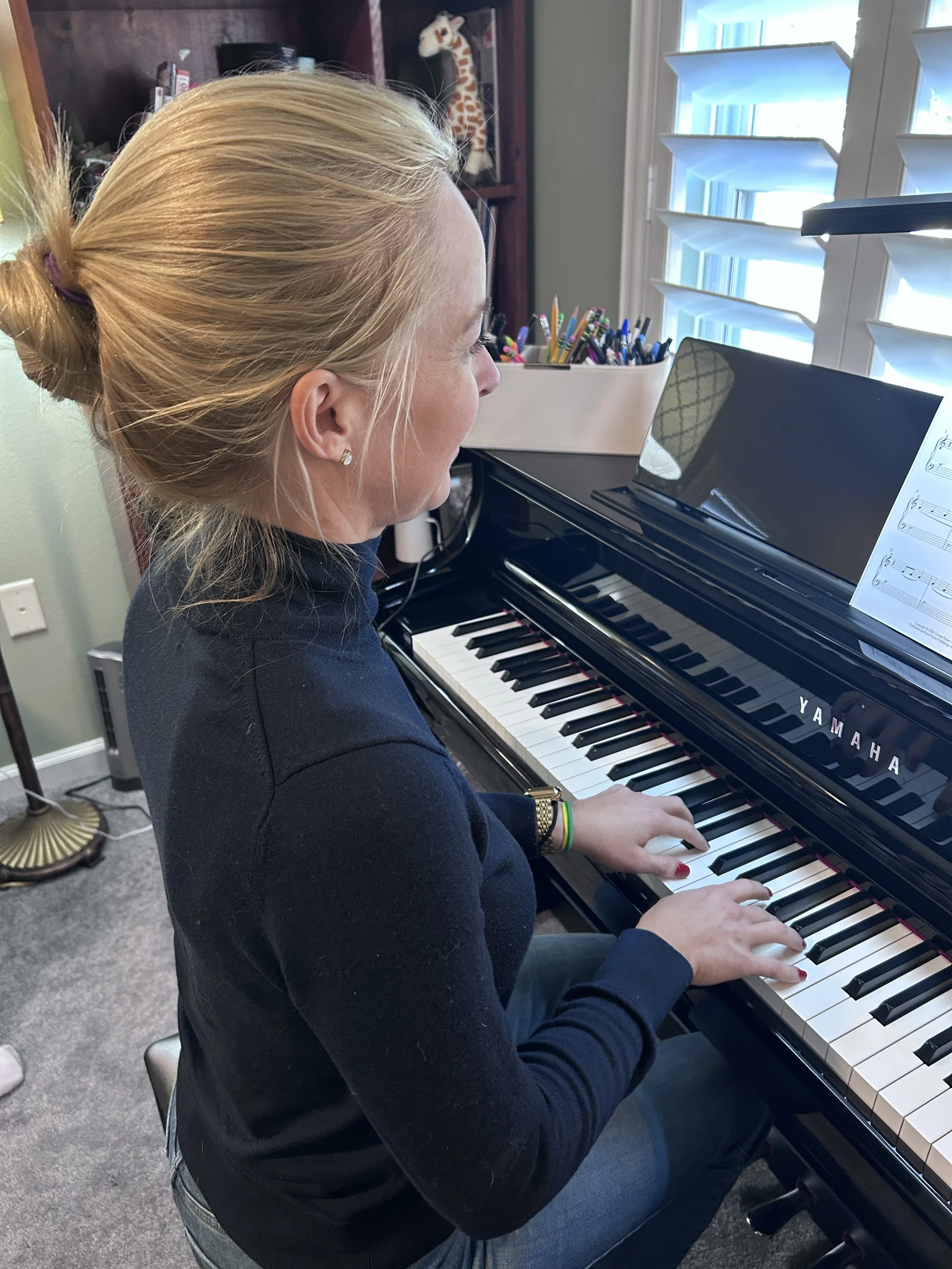 A woman with blonde hair tied in a bun playing a Yamaha grand piano in a room with a window, bookshelves, and colorful writing utensils.
