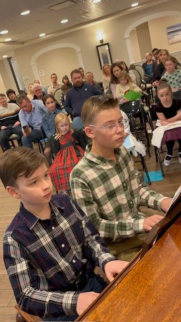 Children and adults seated in a room, with two boys in the foreground playing the piano, surrounded by an audience of various ages.
