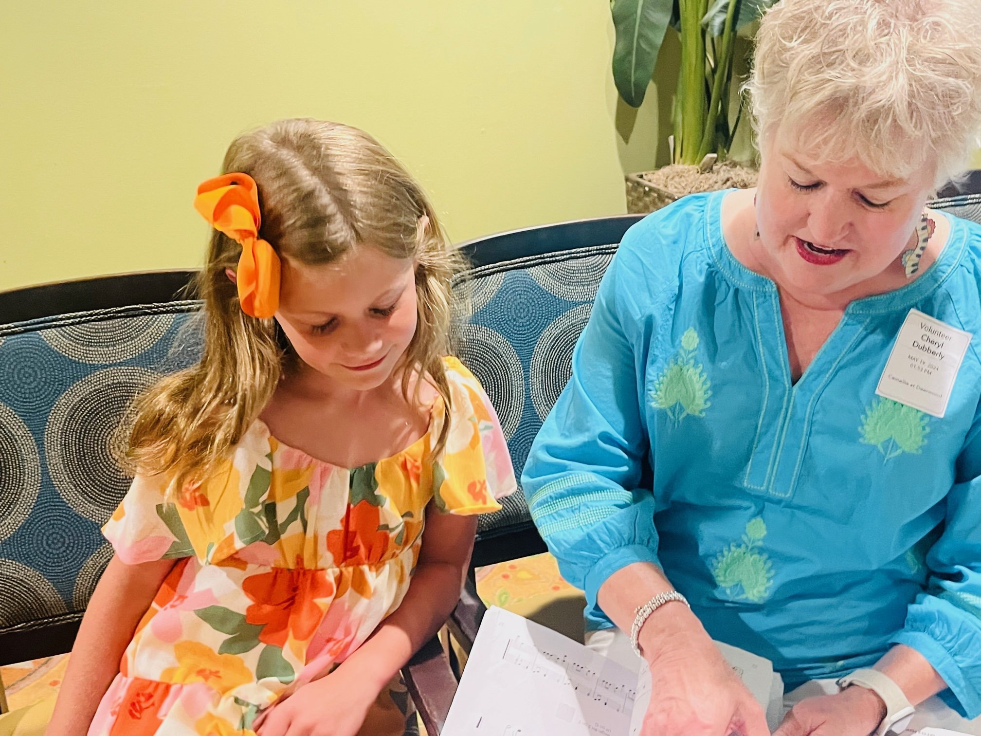 An elderly woman with short white hair, wearing a blue blouse and a name tag, is showing something on a paper to a young girl with long wavy brown hair, wearing a colorful floral dress and an orange bow in her hair, sitting on a patterned blue and black chair.