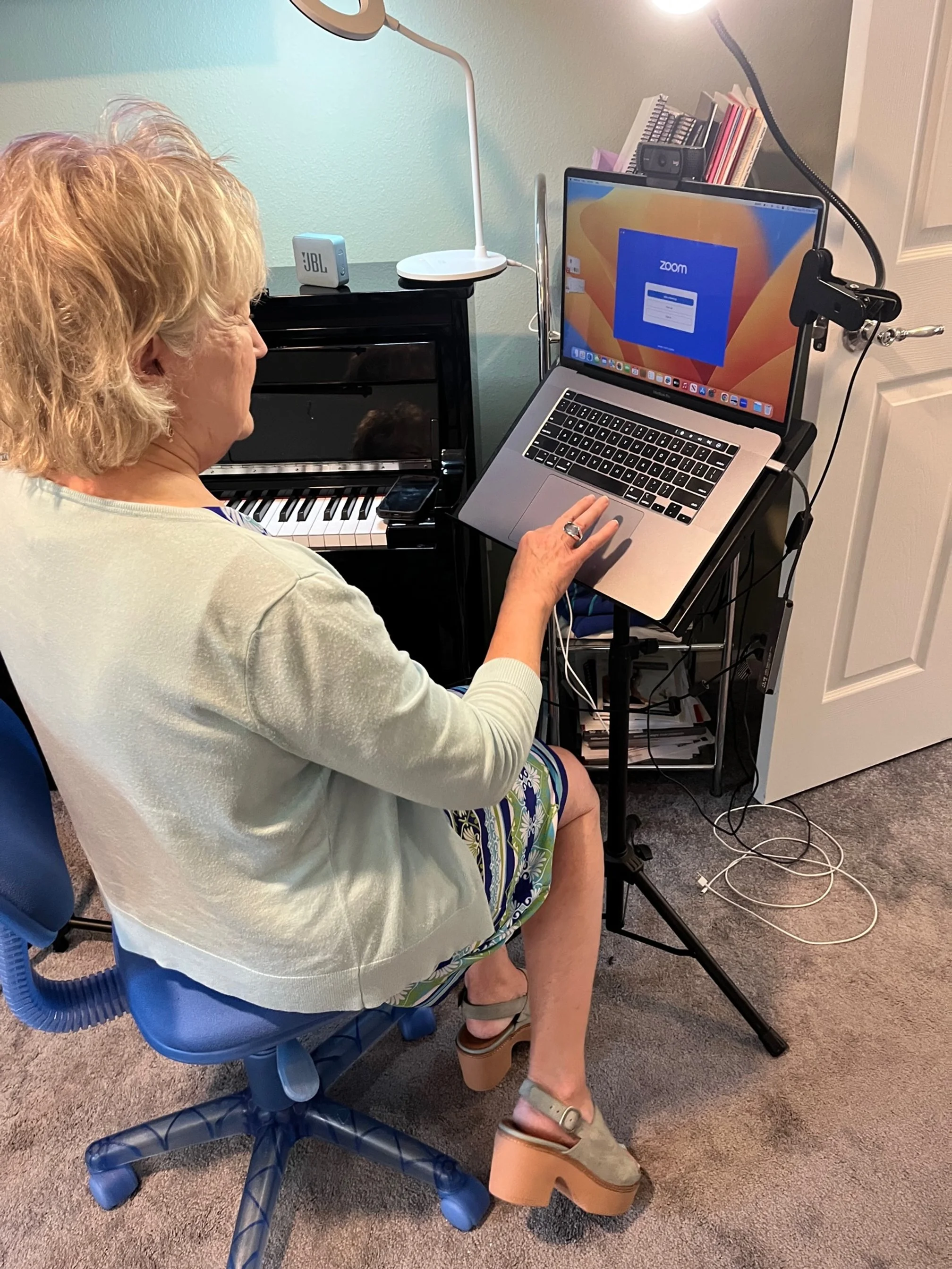A woman with blonde hair sitting on a blue rolling chair, using a laptop on a stand. Behind her, there is a small black piano with a smartphone resting on it, a white desk lamp, and a blue speaker. The woman appears to be participating in a Zoom call. The workspace is cluttered with various items including cables and papers.