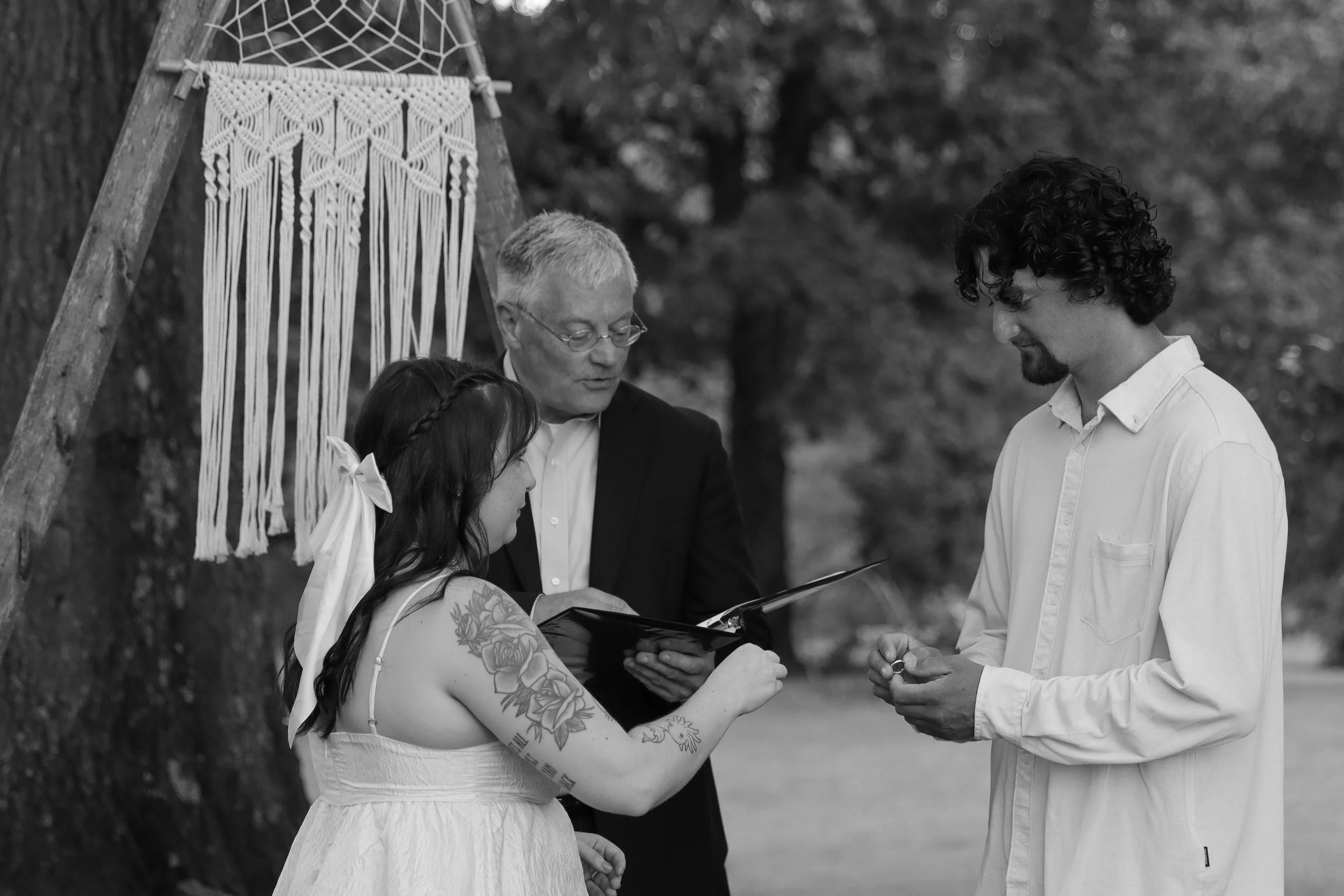 Bride and groom walking down an outdoor aisle, decorated with pampas grass and flowers. The bride is in a white lace dress holding a bouquet, and the groom is in a burgundy velvet jacket with a bow tie.