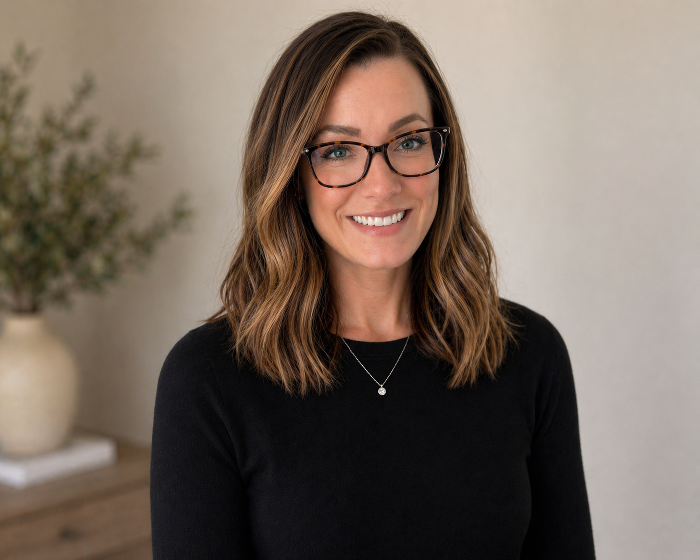 A woman with shoulder-length light brown hair, wearing black-framed glasses, earrings, and a sleeveless top, smiling at the camera with a plain white wall in the background.