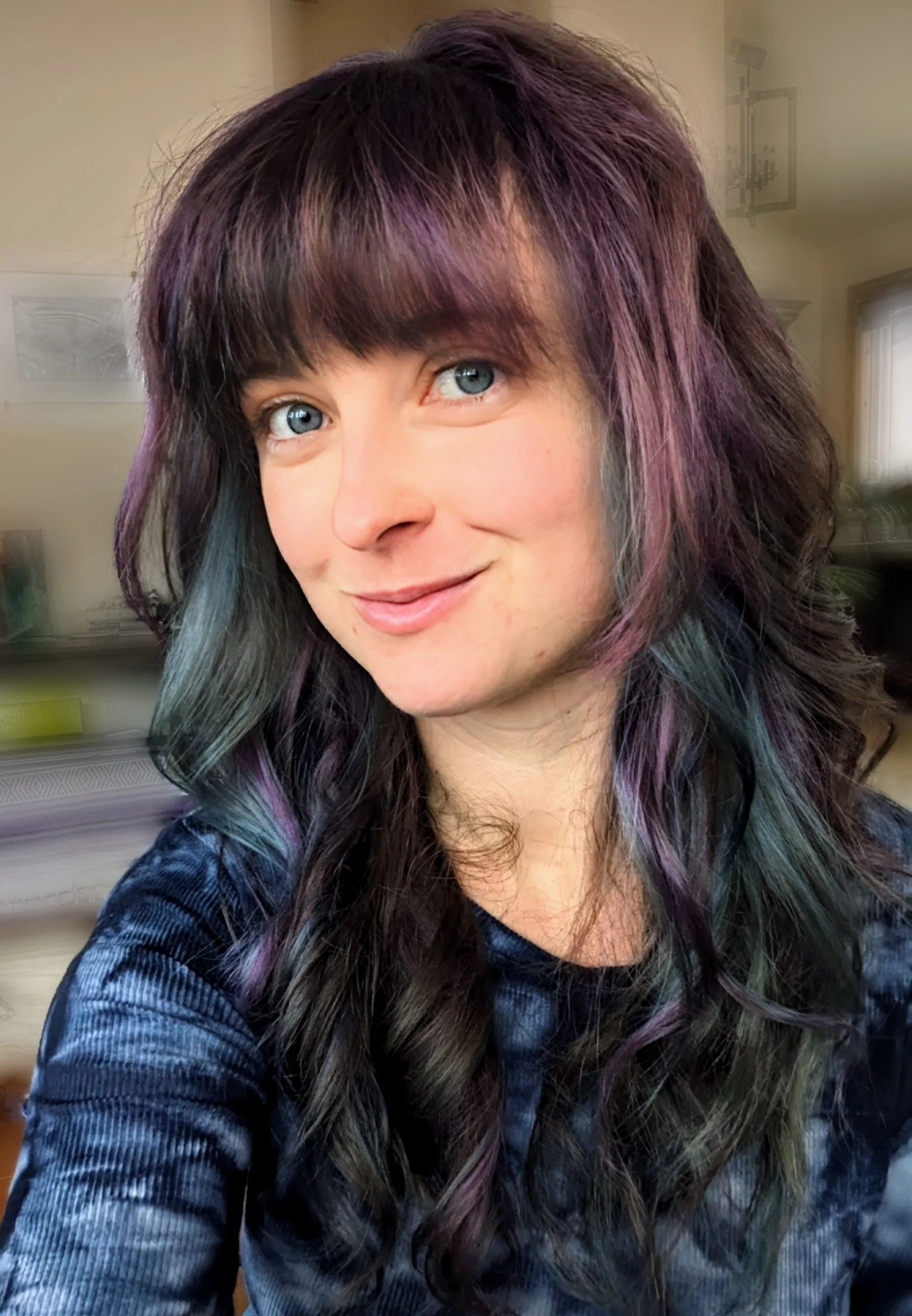 Close-up of a woman with shoulder-length multicolored hair and blue eyes, smiling, indoors with a blurred background.