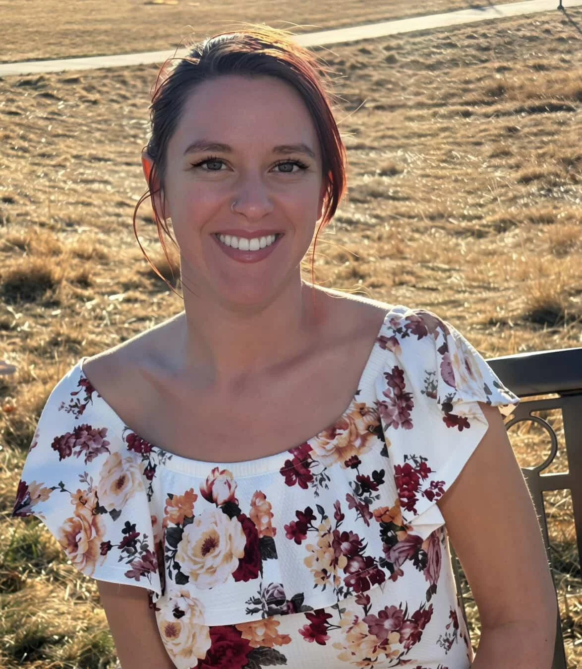 A young woman with short dark red hair, light skin, and a nose piercing, smiling outdoors during sunset, wearing a white floral dress, sitting on a black metal bench.