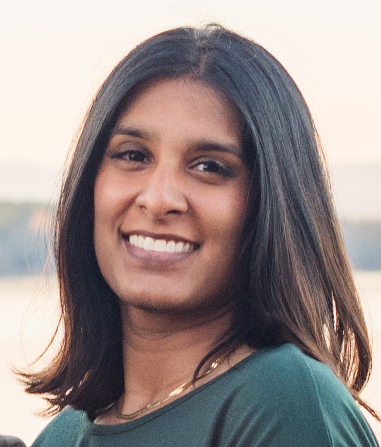 A woman with shoulder-length dark hair smiling outdoors during sunset.