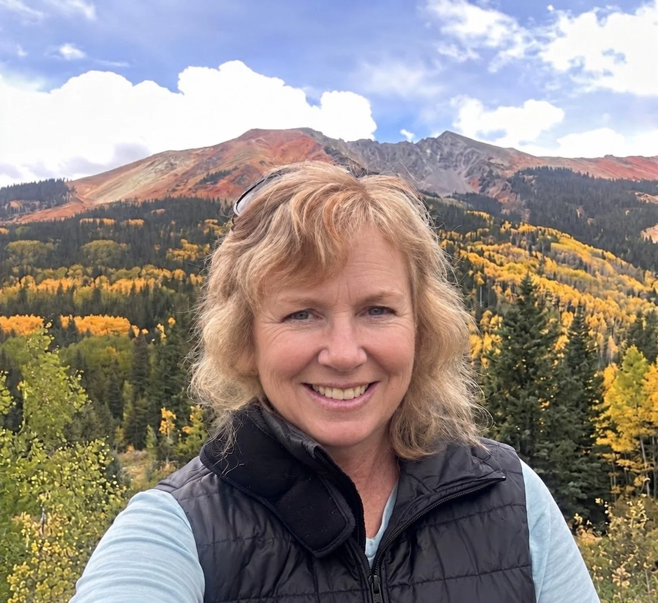 A woman smiling outdoors with a mountain and autumn forest in the background.