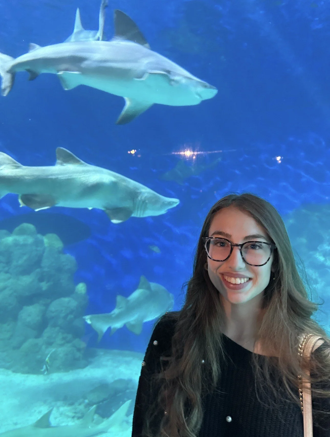 A woman with long brown hair, glasses, and a black top smiling at the camera, standing in front of an underwater aquarium with sharks swimming in the background.