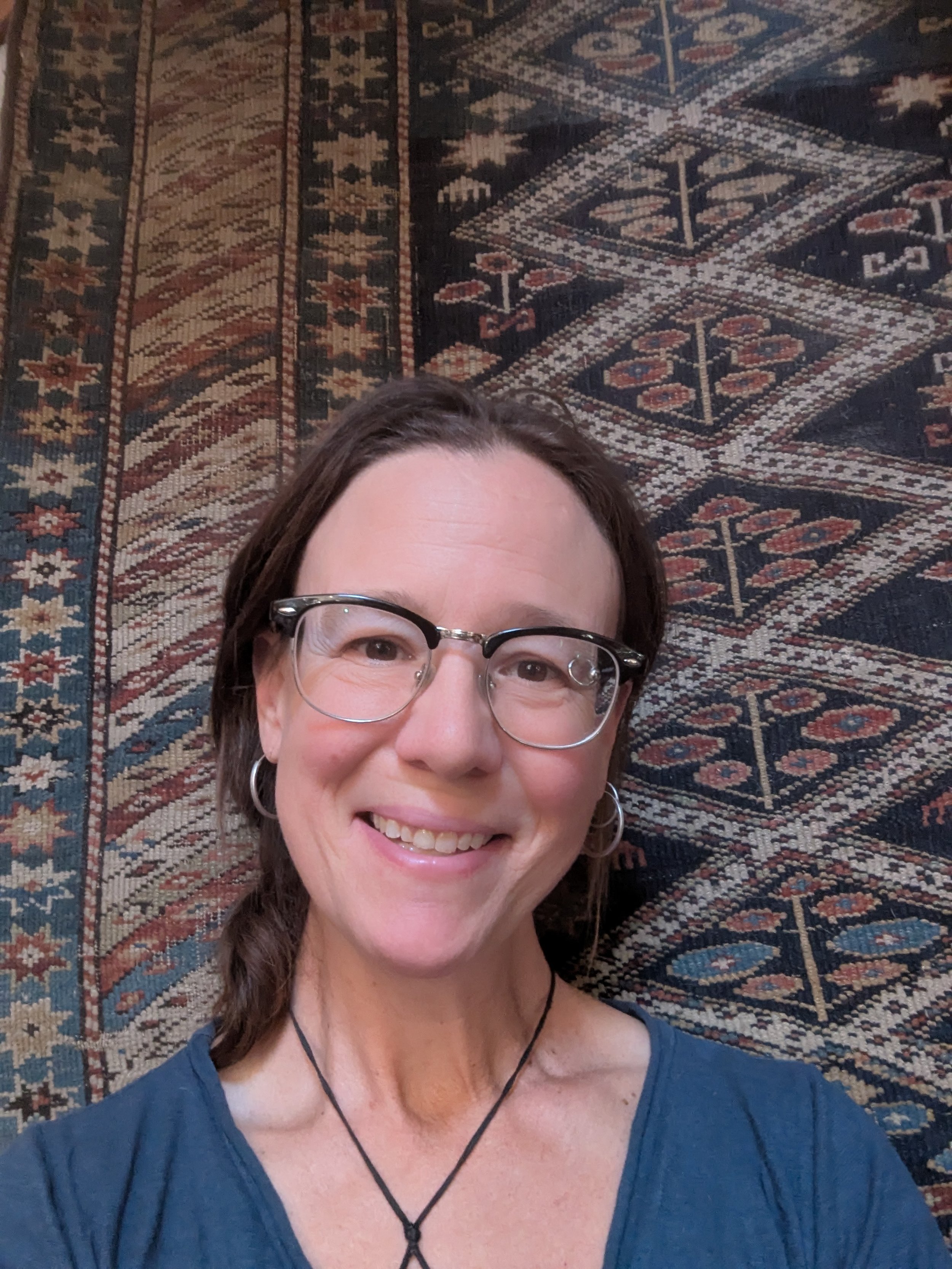 Close-up of a woman with brown hair, glasses, and hoop earrings, smiling, wearing a blue top, with a patterned rug background.