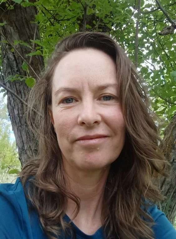 A woman with wavy brown hair and light skin, smiling slightly, standing outdoors near a tree with green leaves in the background.