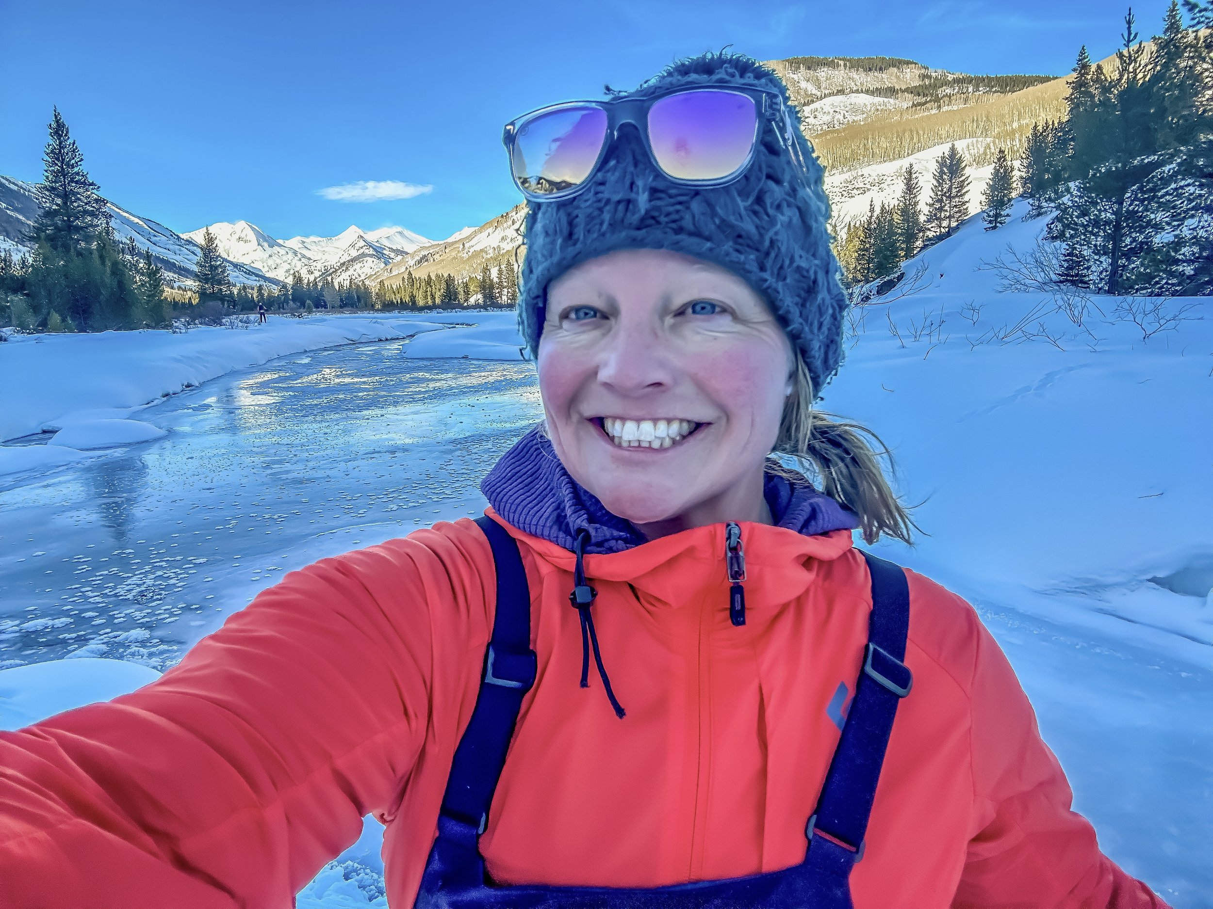 A woman taking a selfie outdoors in a snowy mountainous landscape, wearing a red jacket, a gray knit hat with sunglasses perched on top, and a purple scarf.