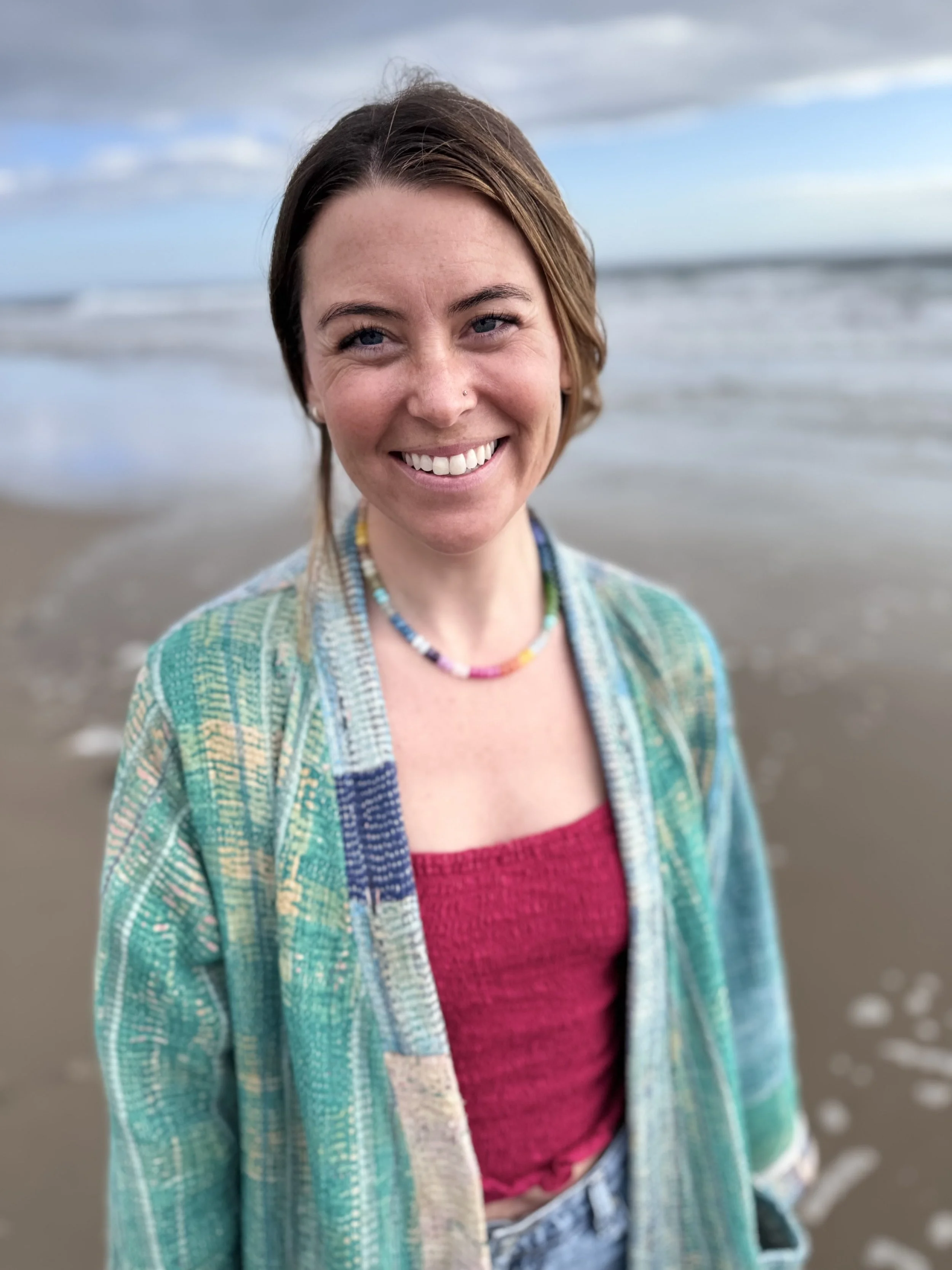 A woman smiling at the camera on the beach with ocean waves in the background.