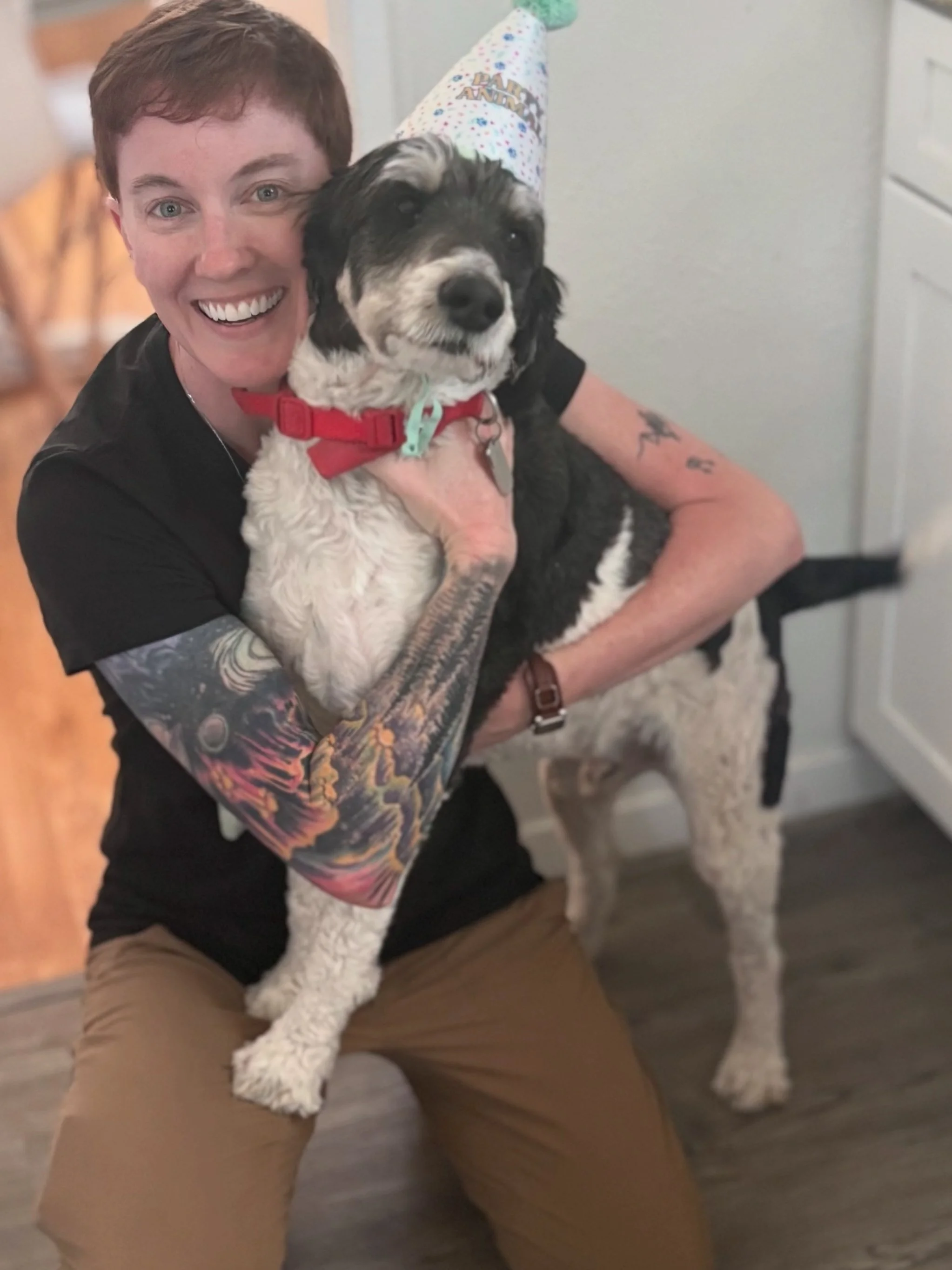 Person wearing a black T-shirt with a red graphic, squatting beside a dog wearing a colorful birthday hat with green fringe. The person has short hair and glasses, smiling at the camera inside a room with wooden flooring.