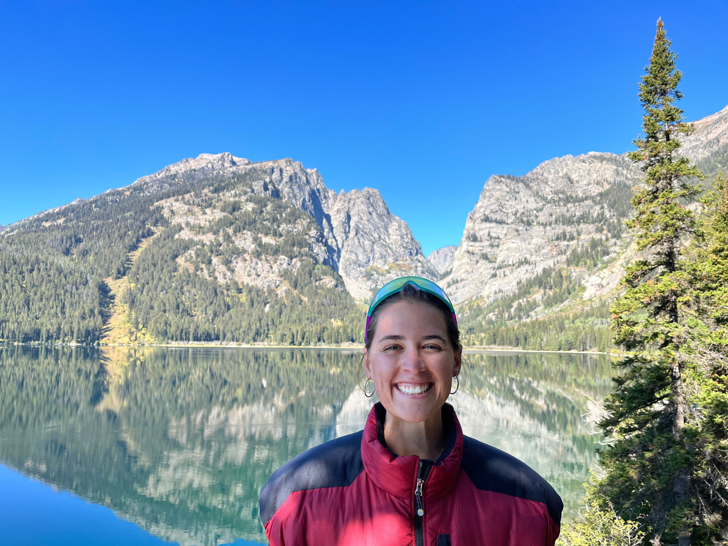 A smiling woman with dark hair, wearing a red and black jacket and sunglasses, standing in front of a calm lake with mountains and trees in the background.