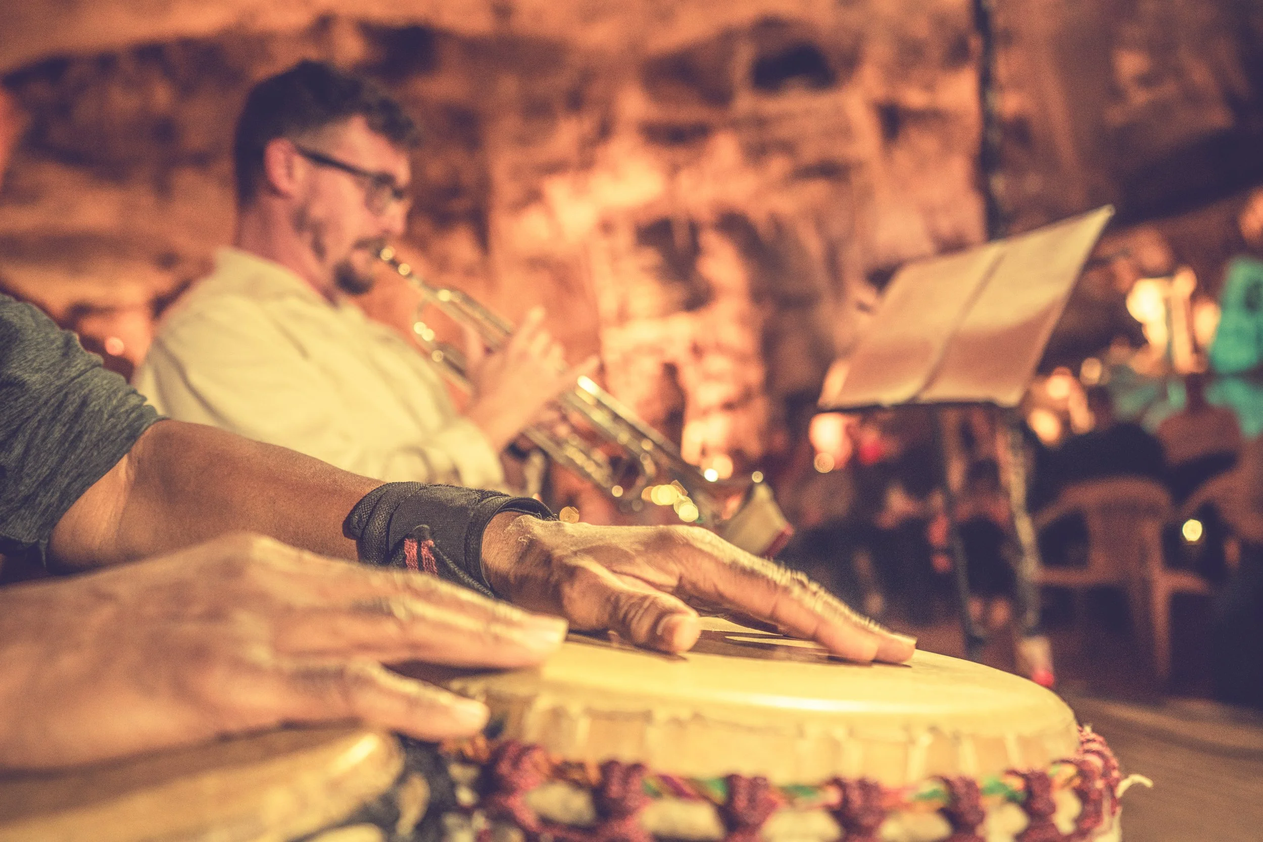 Close-up of hands playing a drum with a musician playing a trumpet in the background at a live music event.