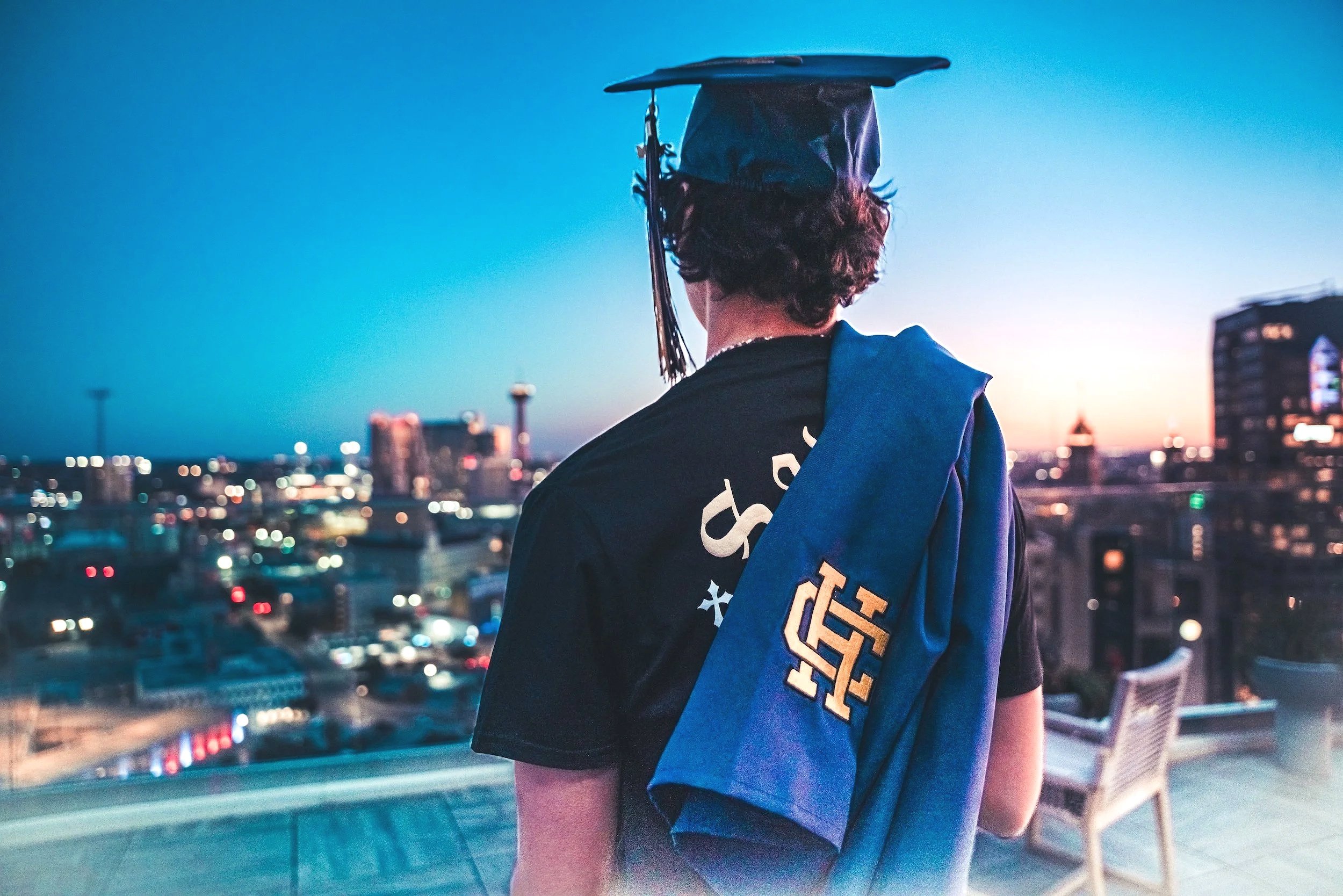 Person with a graduation cap and robe, standing on a rooftop overlooking a city skyline during sunset.