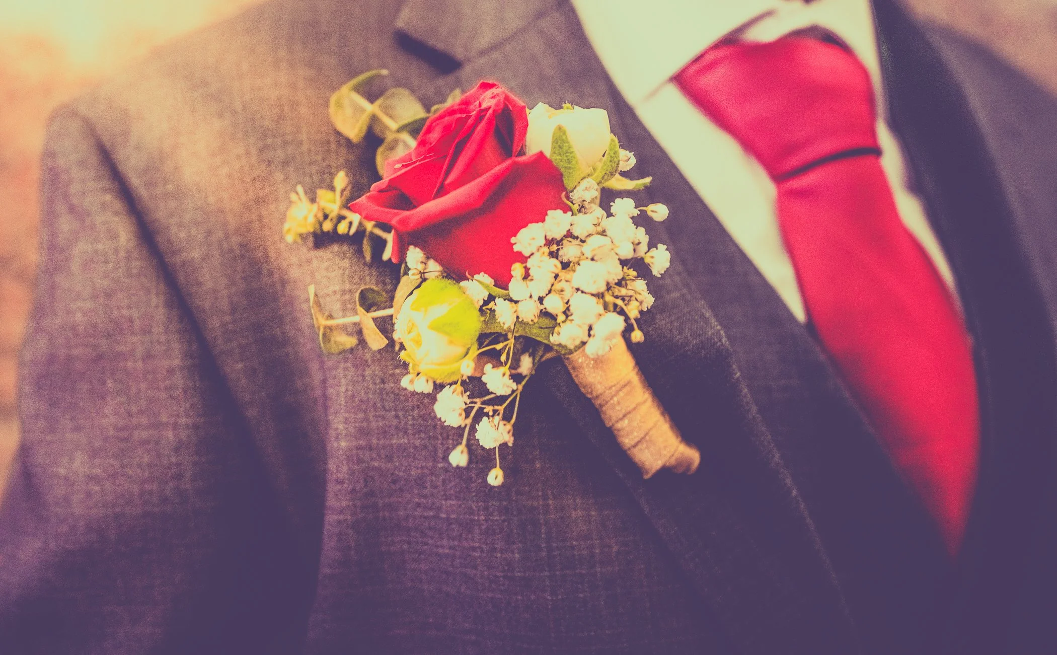 A boutonniere with a red rose, white buds, and greenery, pinned to a gray suit jacket worn with a white dress shirt and red tie.