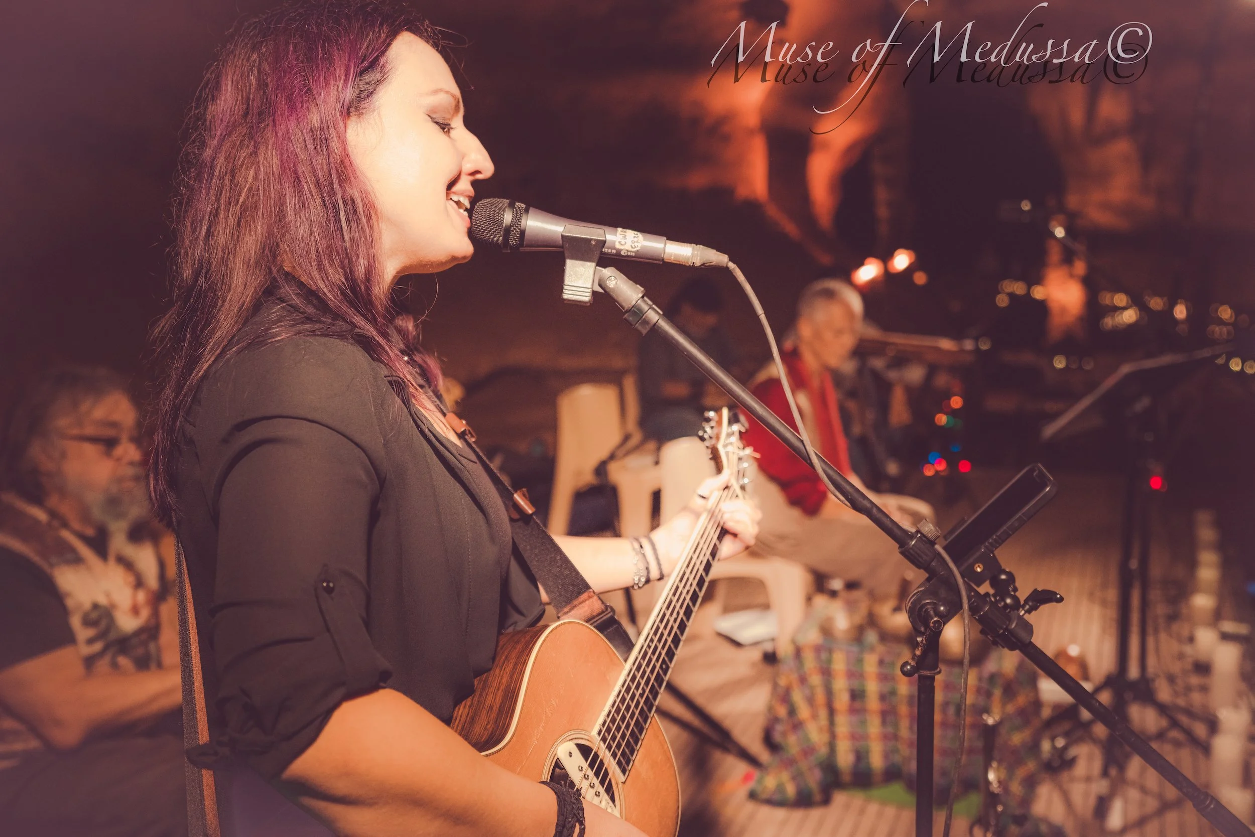 A woman with long purple hair playing an acoustic guitar and singing into a microphone on stage with a dark background. Three other musicians are blurred in the background.