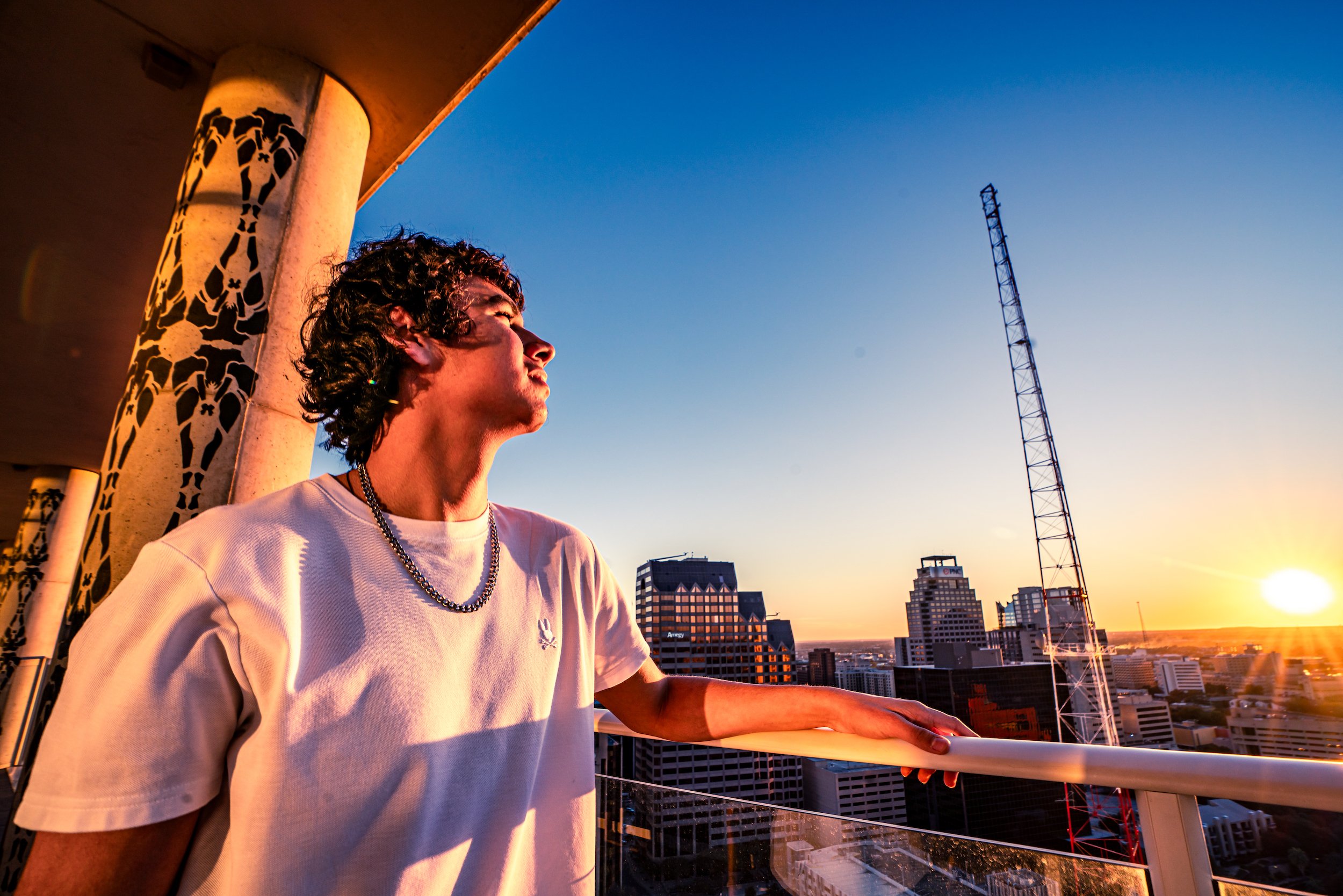 A young man with curly hair wearing a white t-shirt and a silver chain necklace standing on a balcony at sunset, overlooking a city skyline with tall buildings and a radio tower.