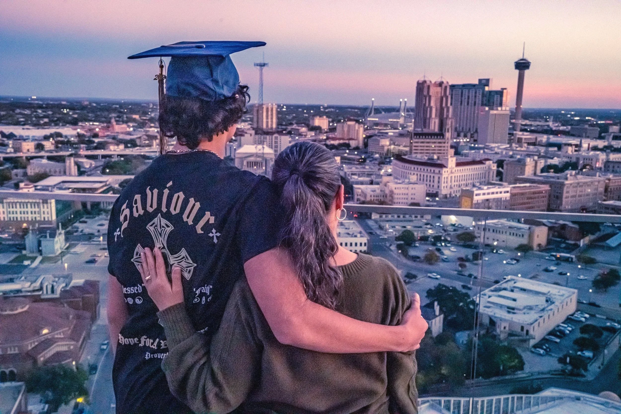 A couple hugging on a rooftop overlooking a city skyline at sunset, with tall buildings and a tower visible in the distance.
