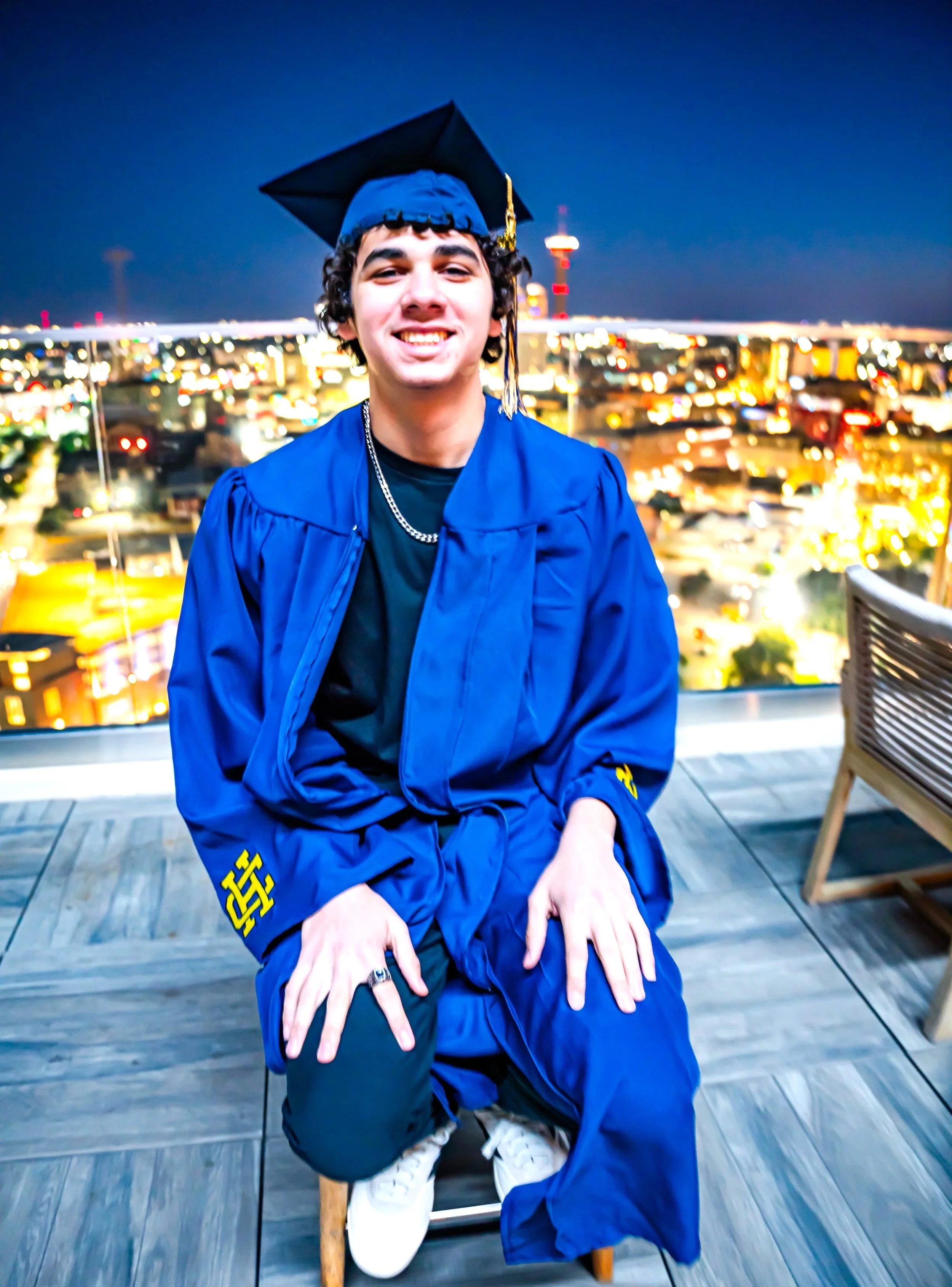 Young man in blue graduation gown and cap, kneeling on a chair on a rooftop at night with city lights and a tower in the background, smiling at the camera.