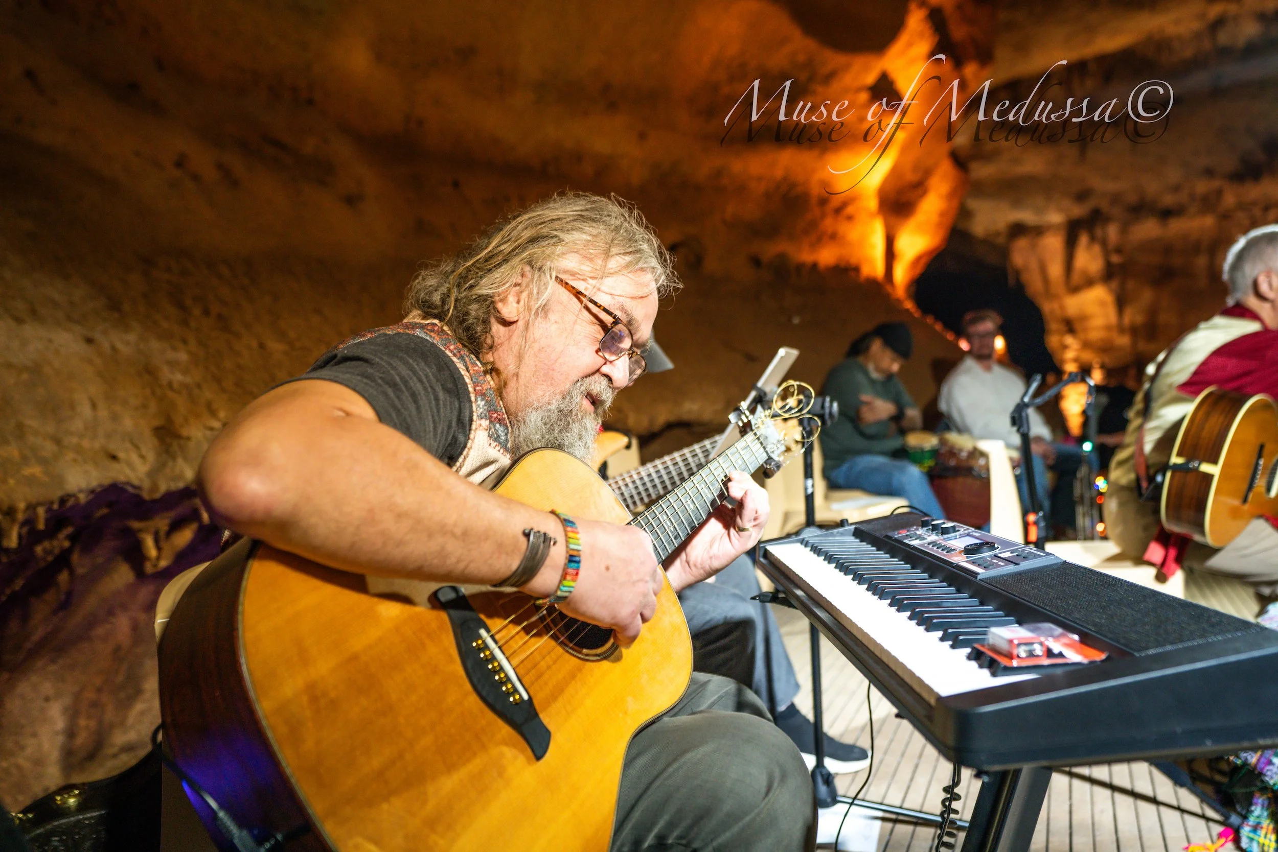 A man with glasses and long gray hair playing an acoustic guitar, sitting on a bench inside a cave with warm lighting. There are other people in the background, some playing instruments and others seated, with a keyboard in front of him.
