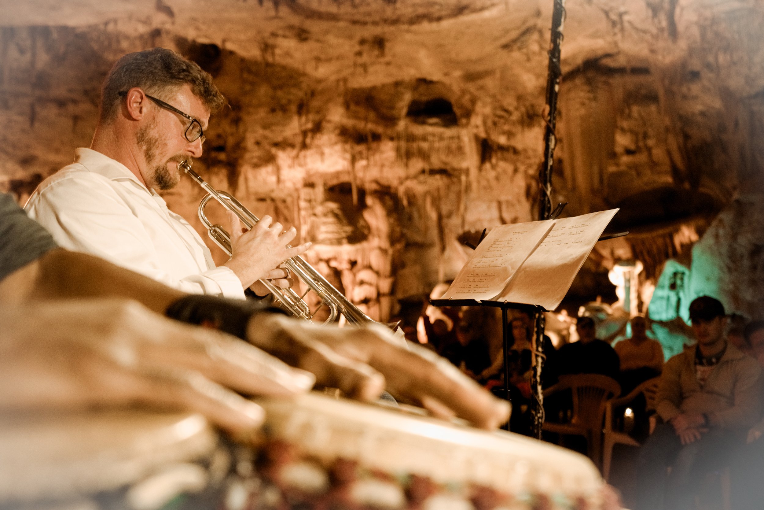 A man with glasses and a beard playing a trumpet during a live music performance in an underground cave with an audience watching.