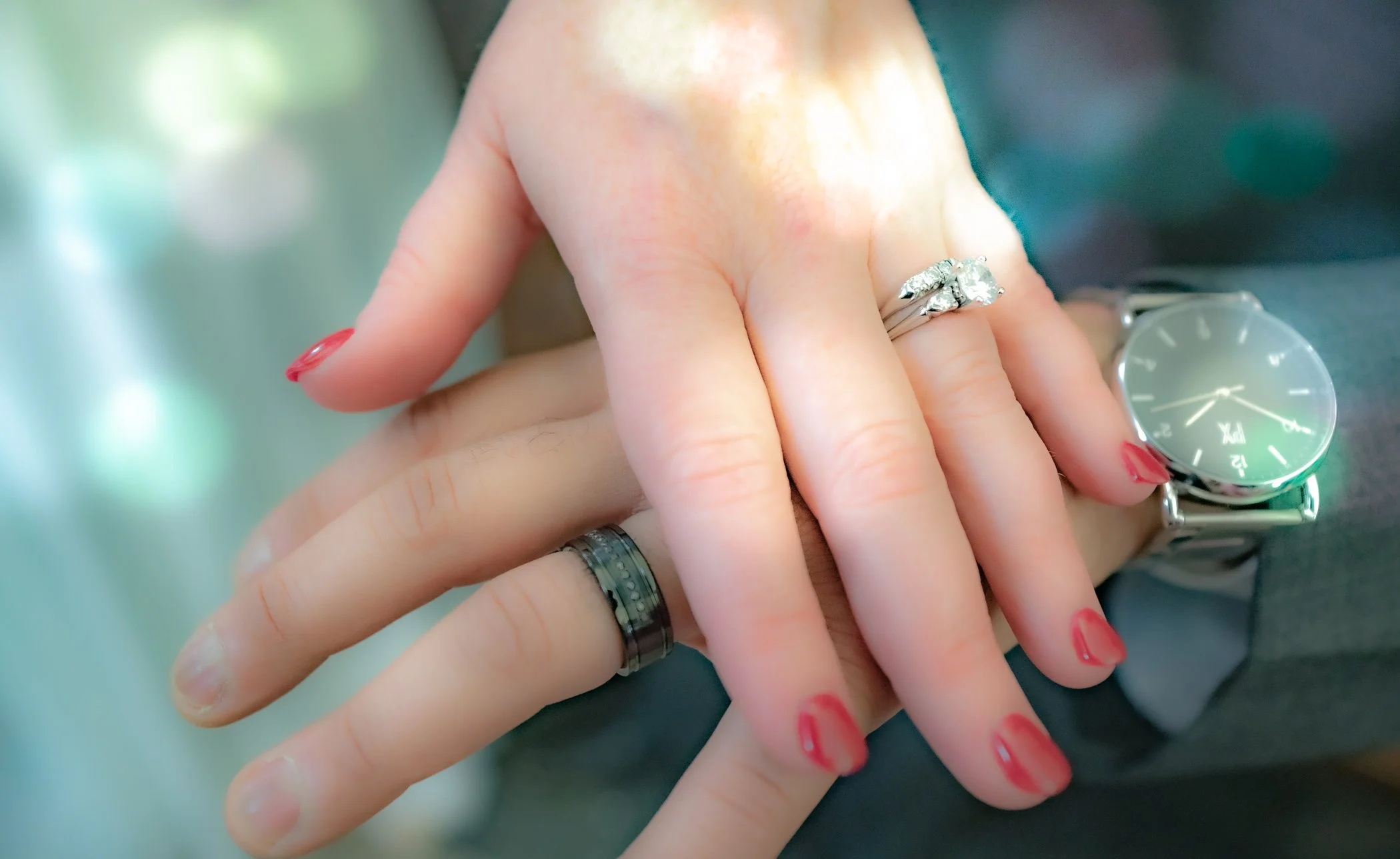 Close-up of two hands with rings, one wearing a watch, resting on a dark fabric surface, with a blurred background.