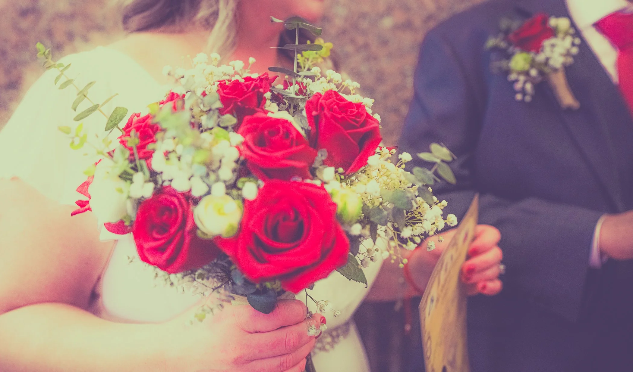 Bride holding a bouquet of red roses and white flowers during a wedding ceremony, with groom in a dark suit and red tie standing nearby.