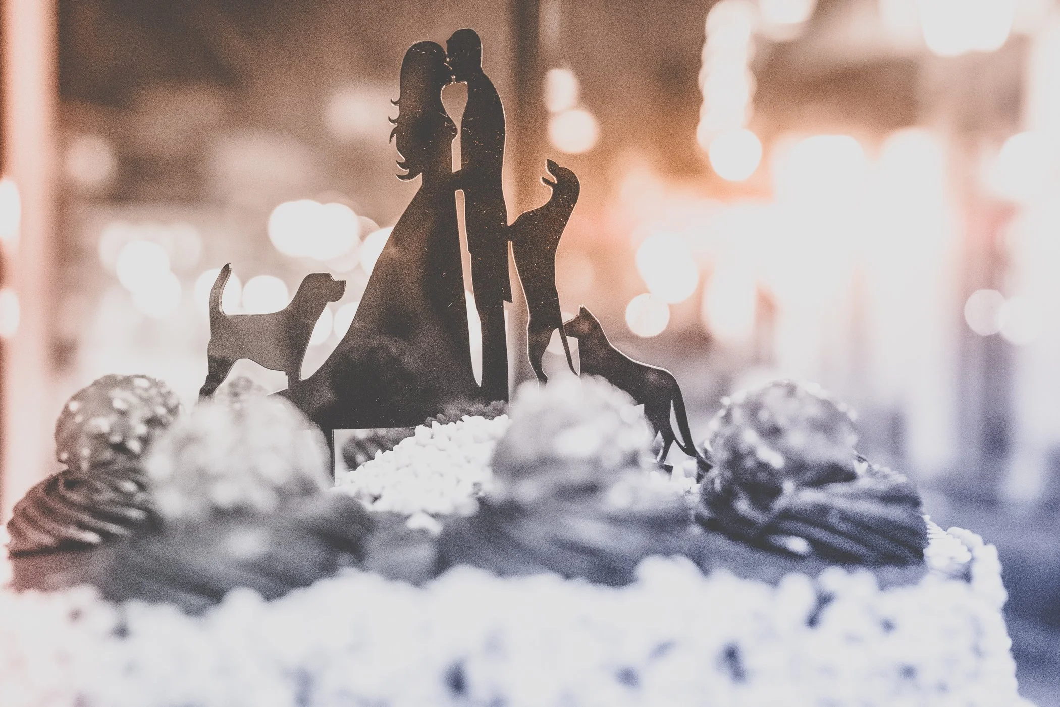 Cake with a silhouette topper of a couple kissing, surrounded by cookies, on a blurred background with warm lighting.