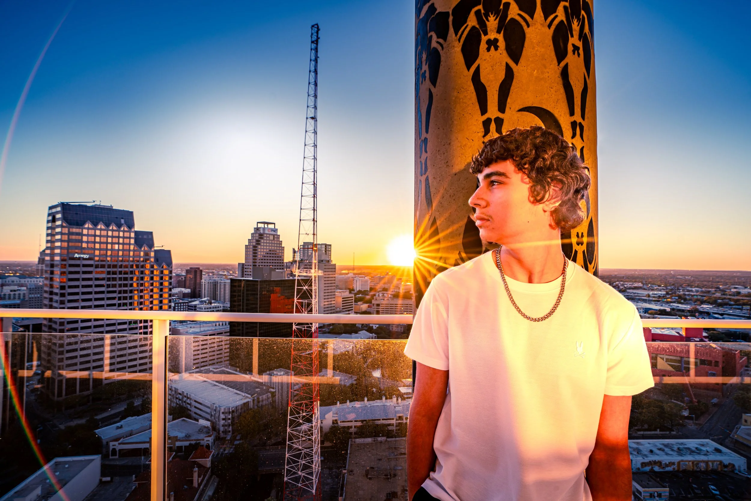 Young man with curly hair wearing a white T-shirt and gold chain standing on a rooftop overlooking a city skyline at sunset with a tower in the background.