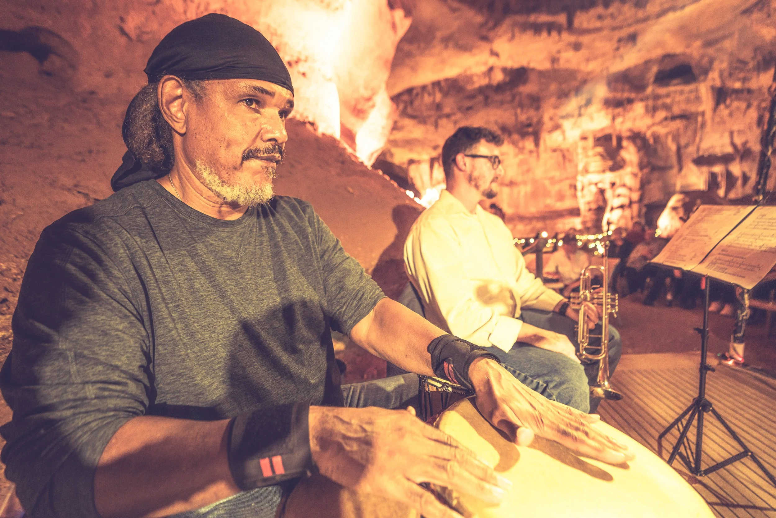 Two musicians playing drums and trumpet in a cave with warm lighting and rock formations.
