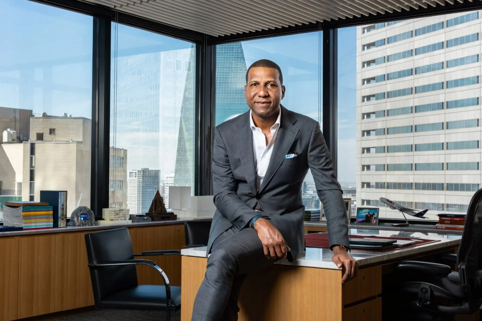 Executive portrait of a man in a dark suit seated beside floor-to-ceiling office windows.