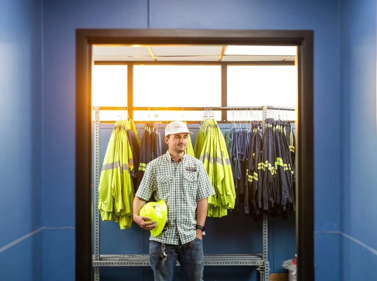 Workplace portrait of a man wearing a hard hat and holding safety gear beside a rack of high-visibility jackets.